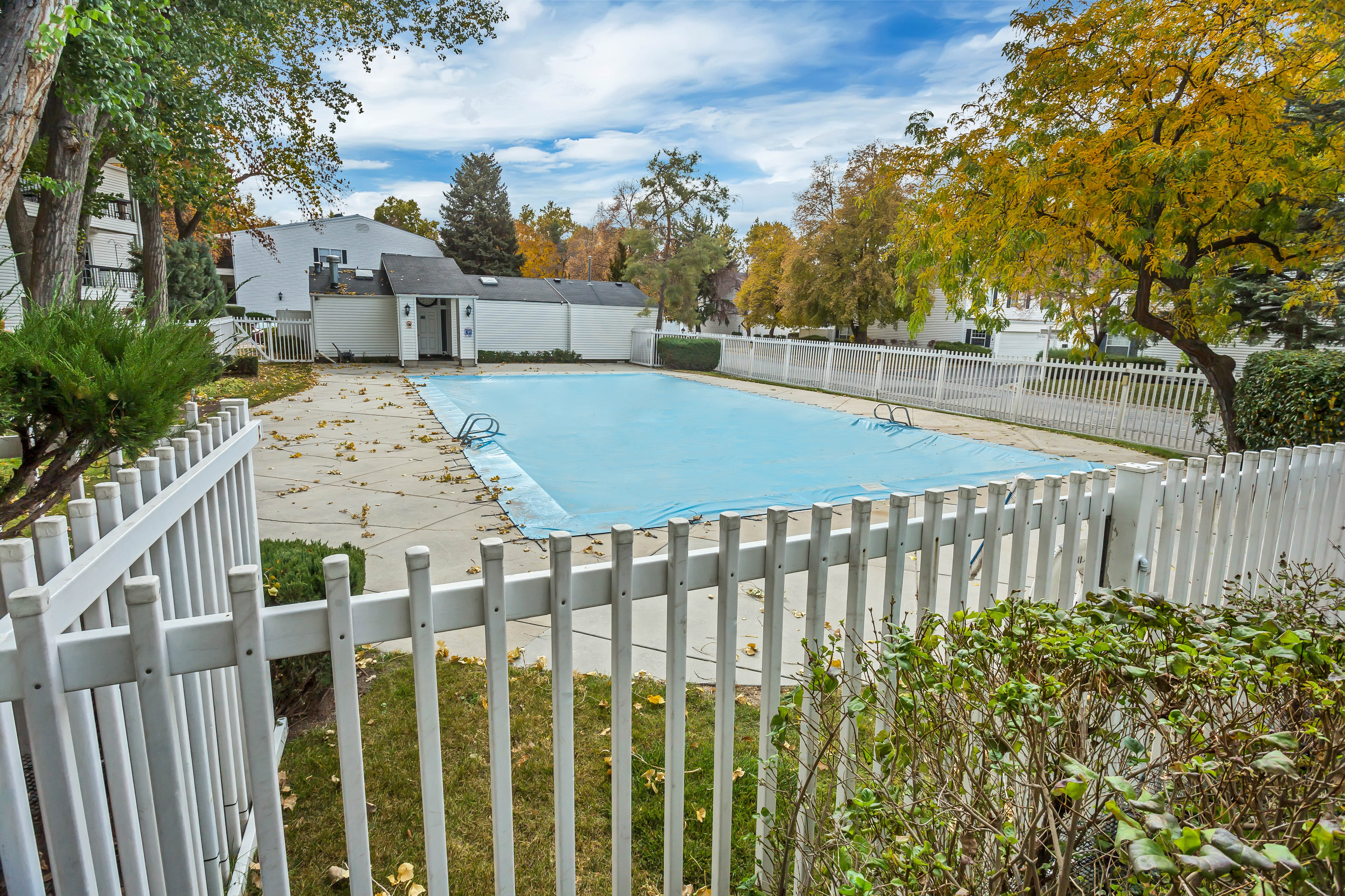 Gated pool area featuring a covered swimming pool, concrete surround, white picket fencing, and mature perimeter trees
