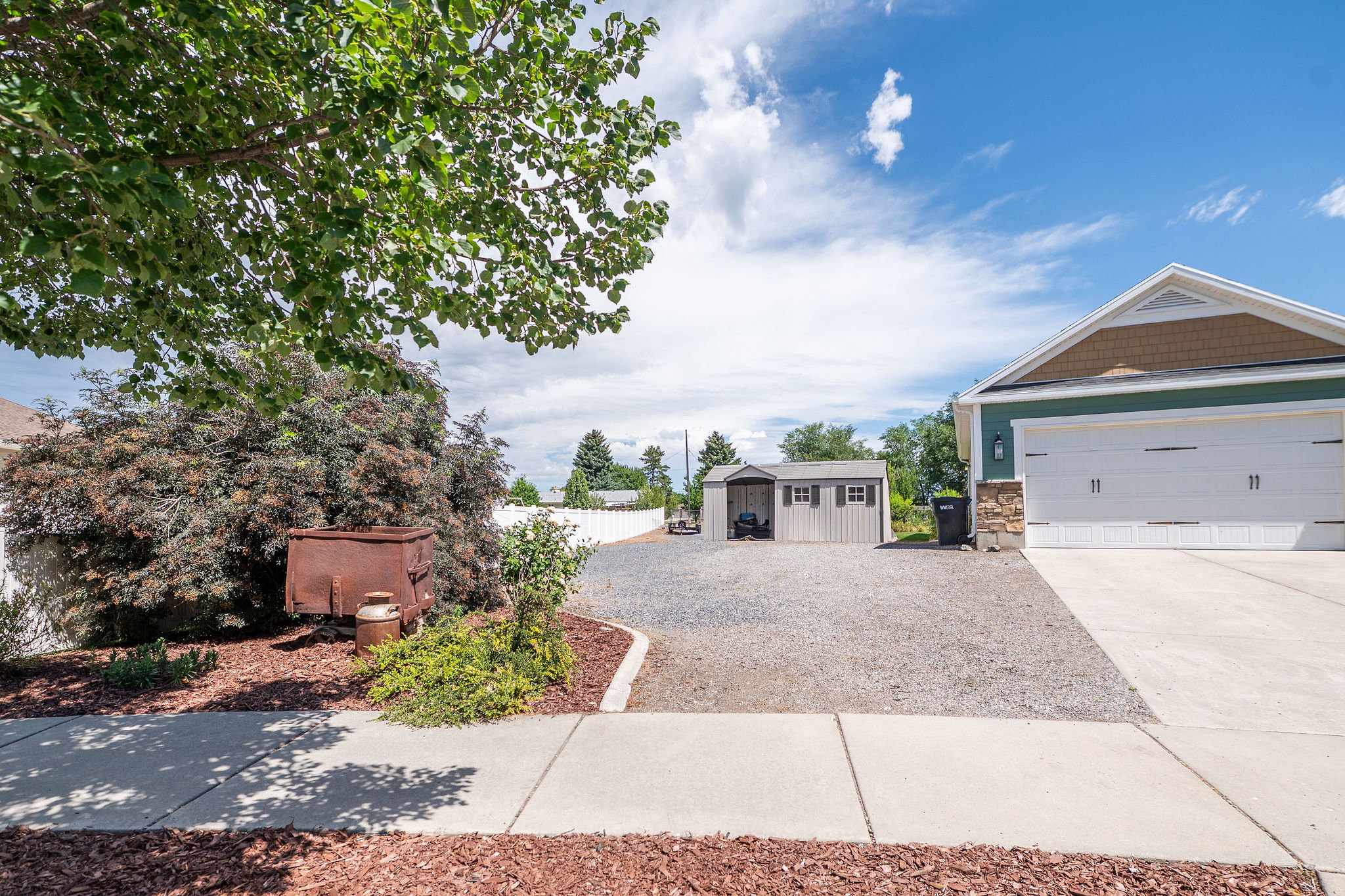 Front exterior showcasing a two-car garage with a concrete driveway, a separate gravel parking area, and two storage sheds