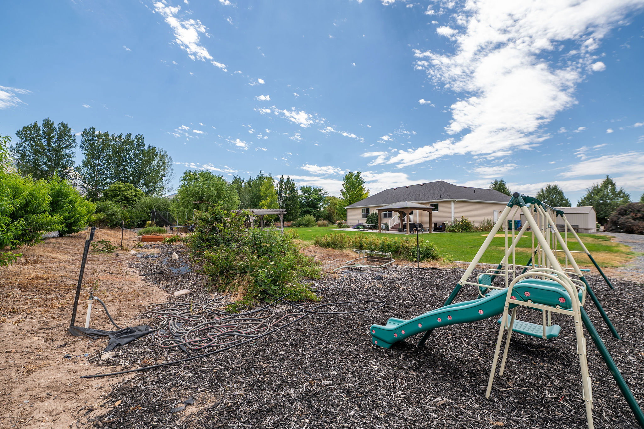 Expansive backyard featuring a designated play area with a slide and swing set, a large grassy area, a covered patio, and mature trees