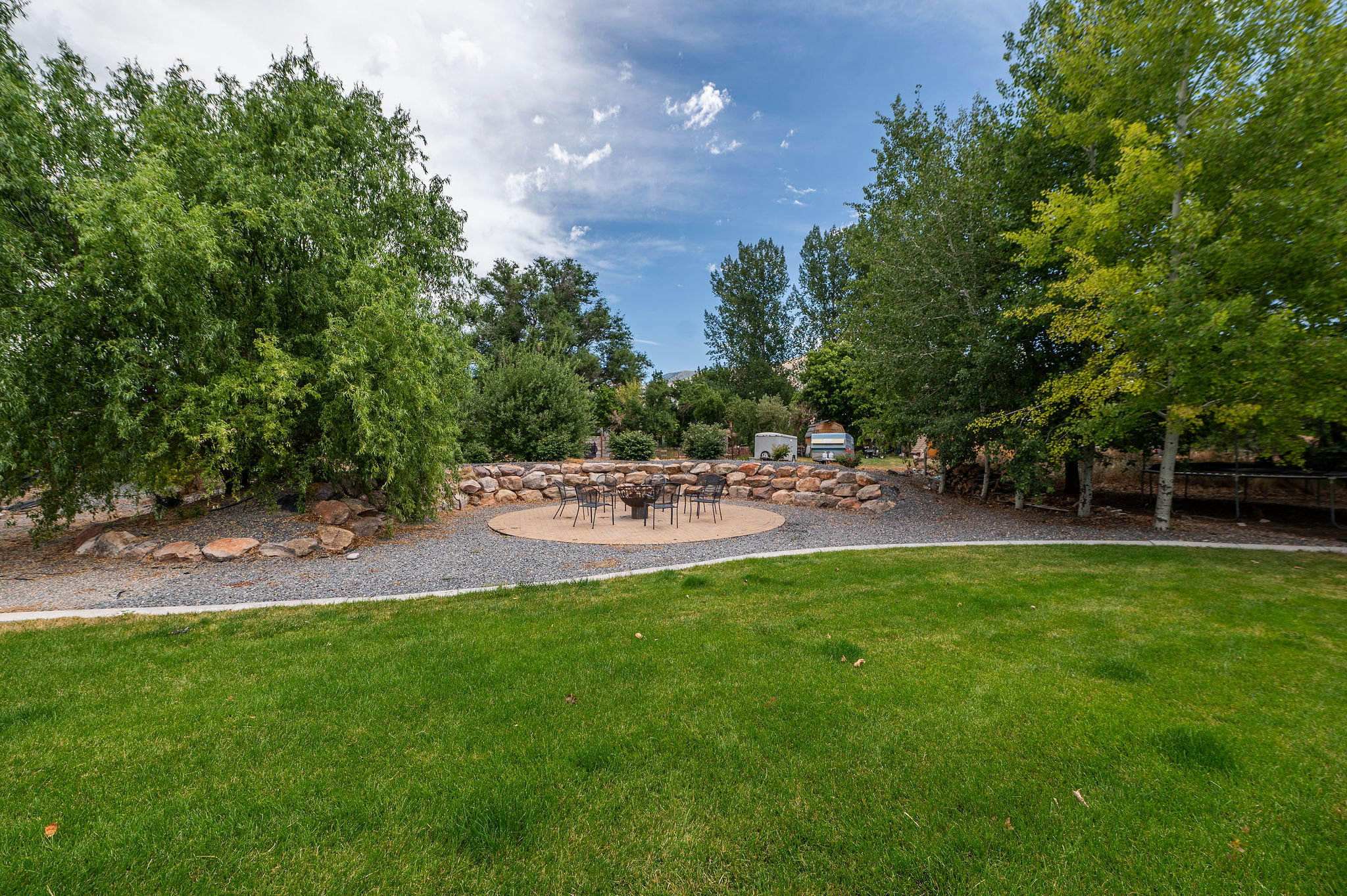 Expansive lawn area featuring a circular patio, a stone retaining wall, mature trees, and a gravel pathway