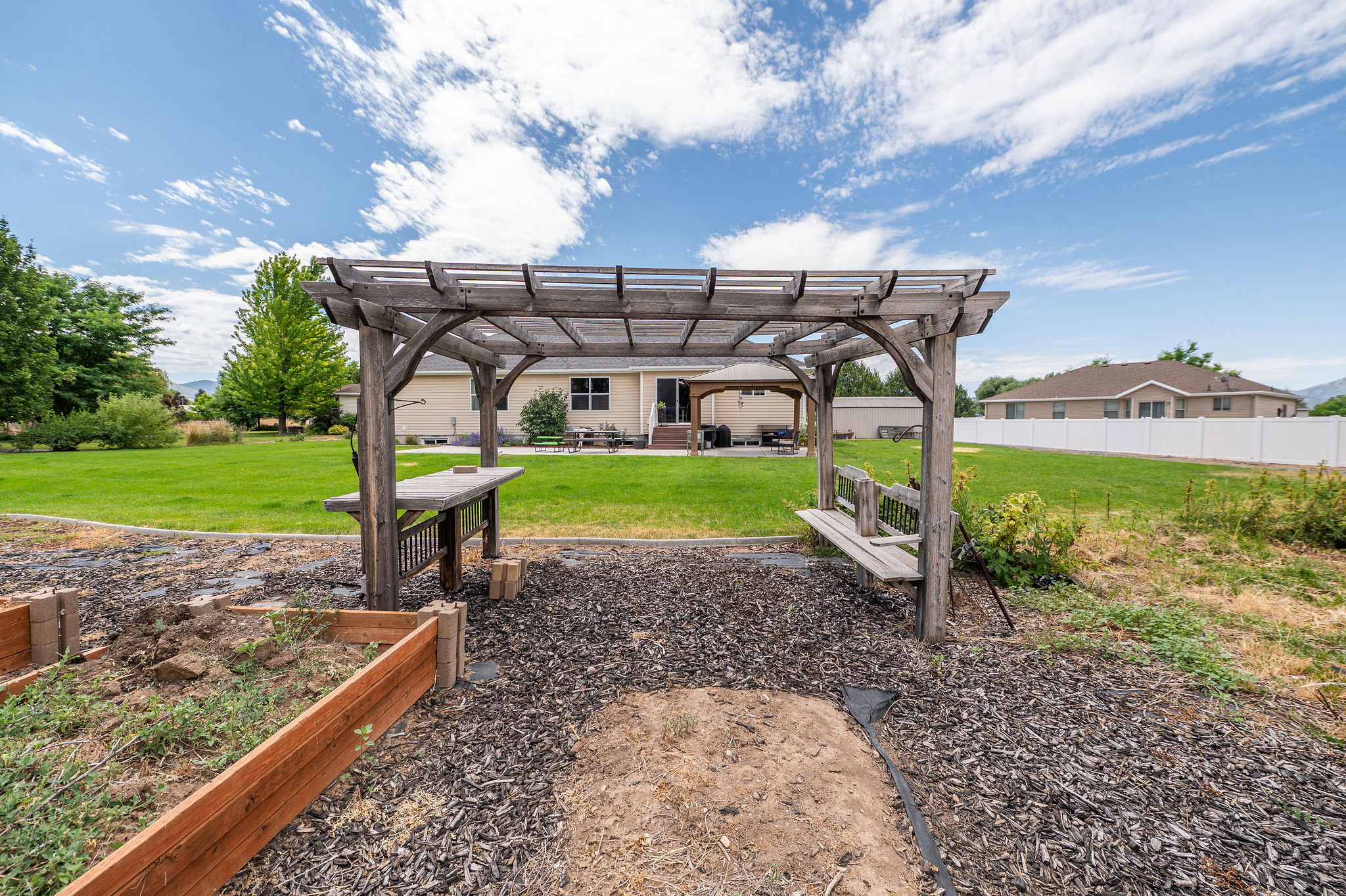Expansive backyard featuring a large wooden pergola with an attached bench, raised garden beds, and a sprawling lawn