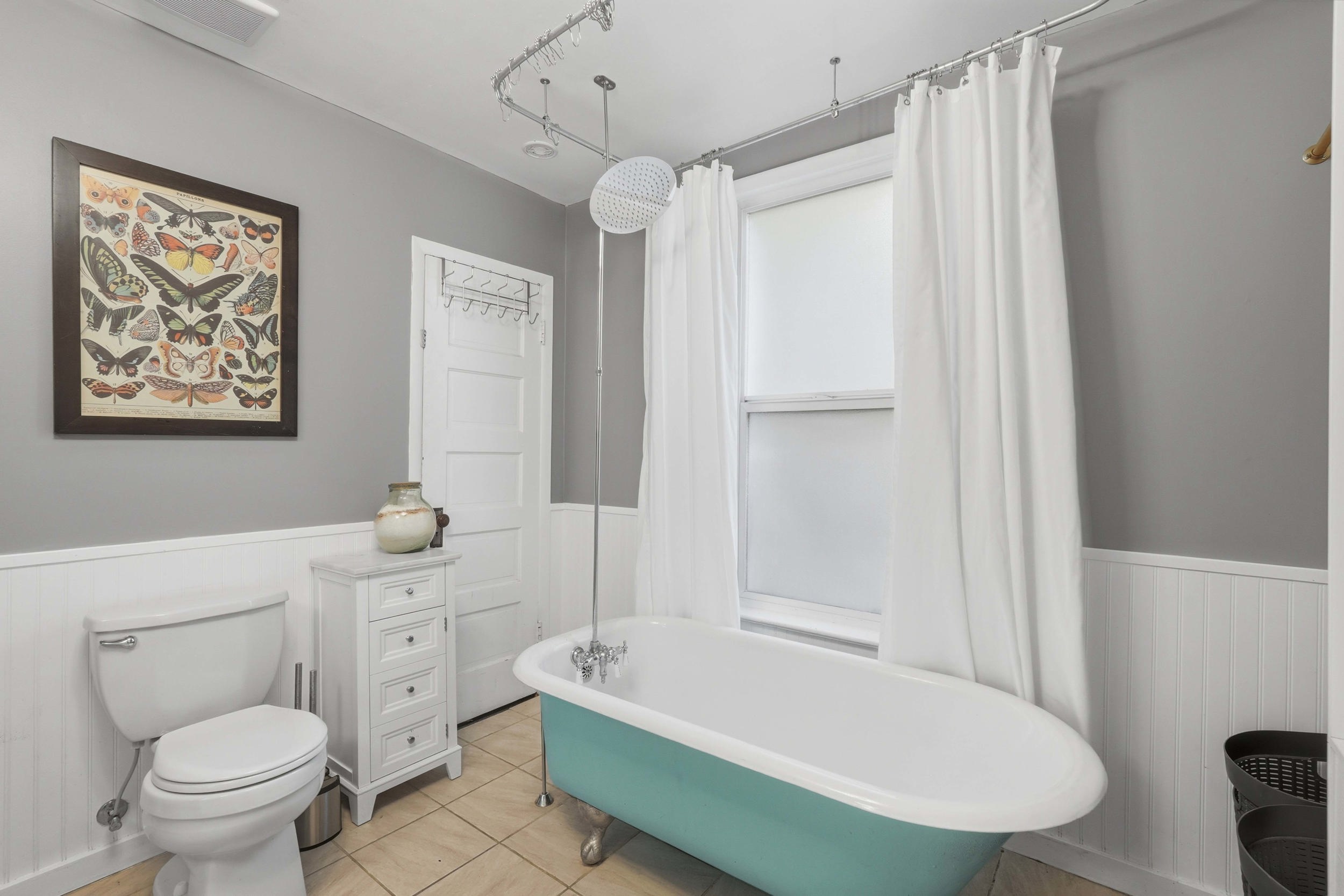 Owners suite. Freestanding clawfoot tub with shower extension, white beadboard wainscoting, light tile flooring, and a white paneled door