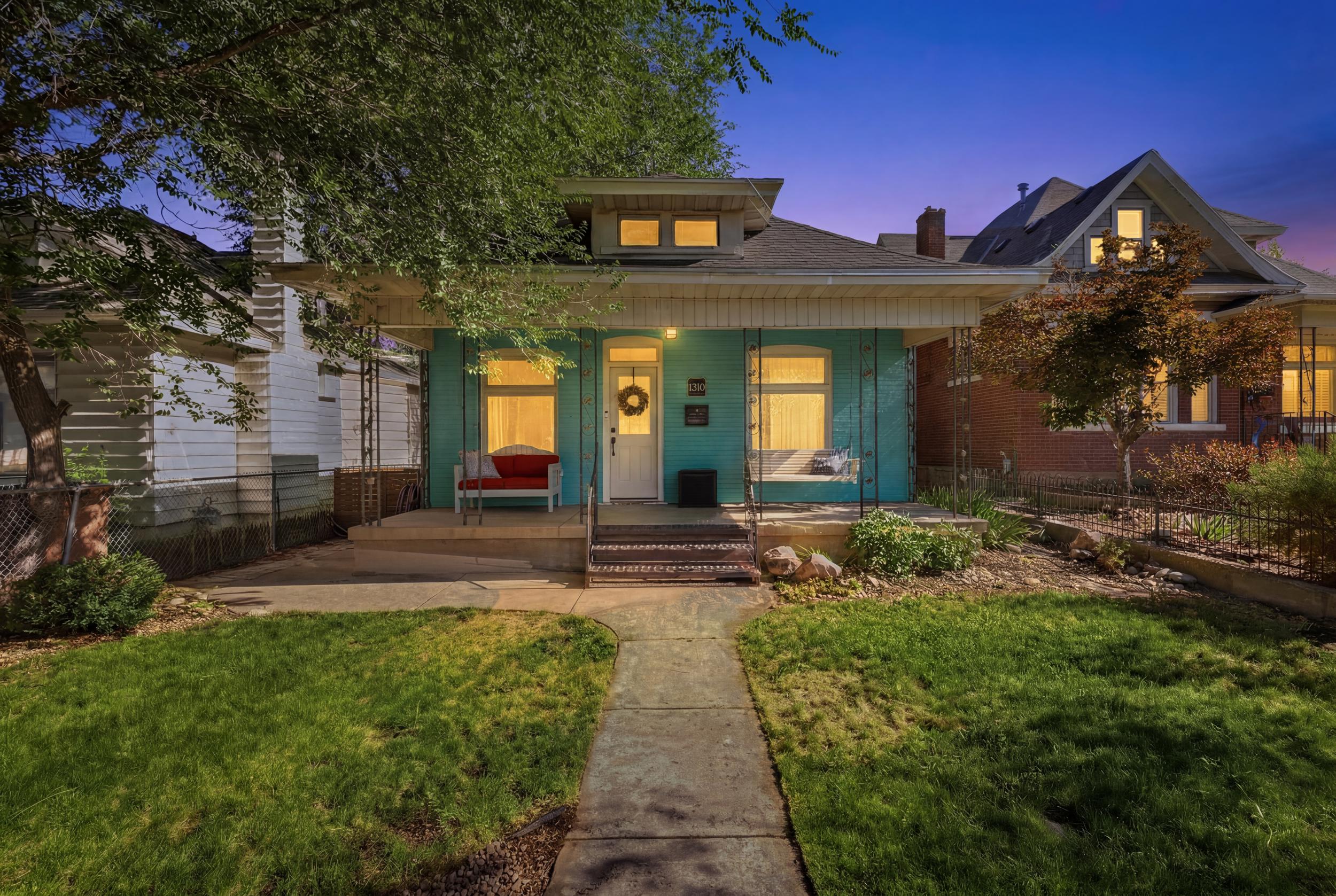 Vibrant front exterior with a light turquoise facade, prominent front porch, mature front yard trees, and a concrete pathway leading to the entrance