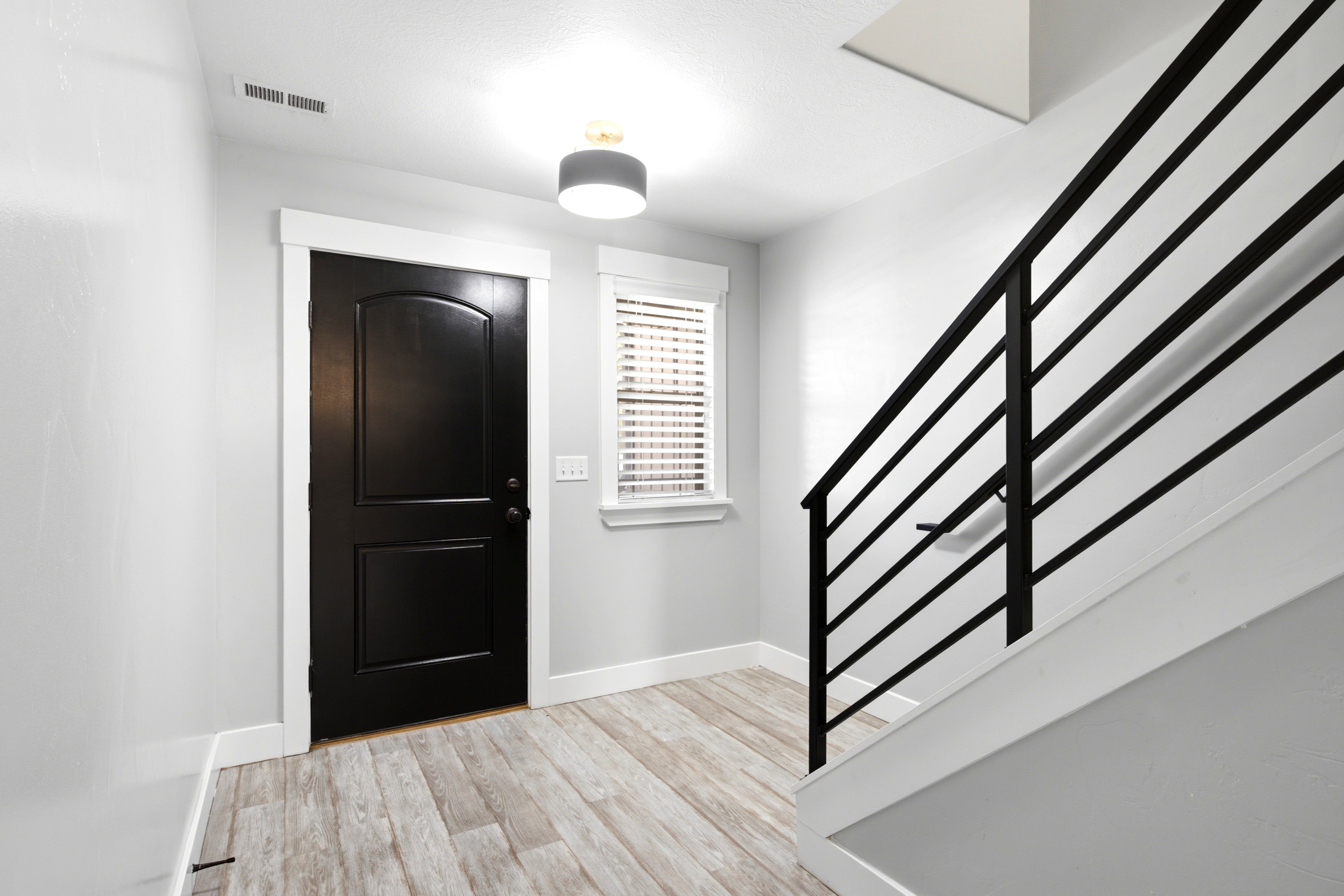 Entryway featuring a solid black door with arch paneling, contemporary flush mount lighting, wood-finish flooring, a window with horizontal blinds, and a staircase with a horizontal bar railing