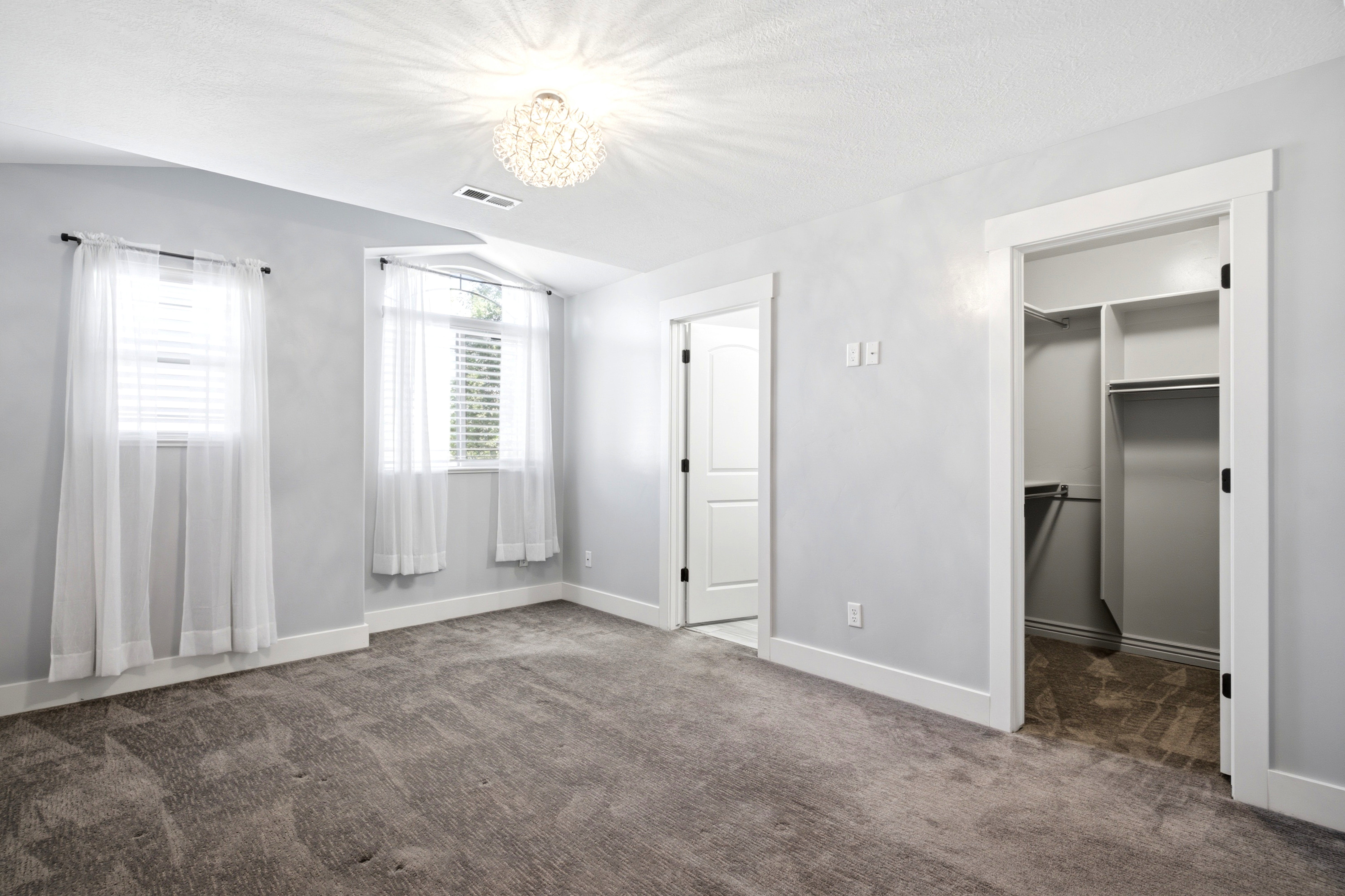 Spacious room featuring neutral carpeting, light gray walls, two windows with blinds, a crystal-style ceiling light fixture, and an open closet with built-in shelving