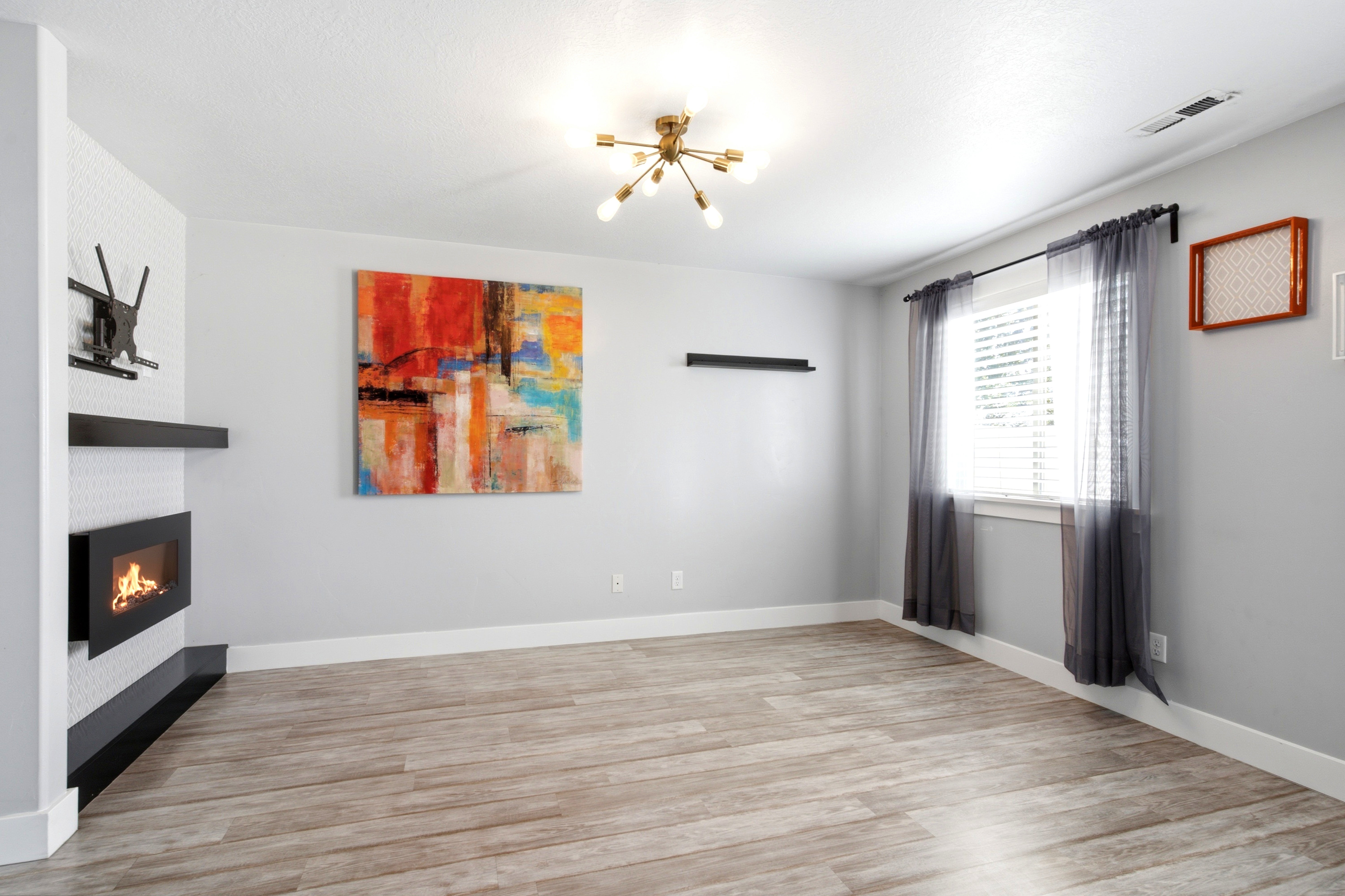 Living space featuring light wood-finish flooring, a modern wall-mounted fireplace with a black mantel, an overhead sputnik chandelier, and a large window with blinds