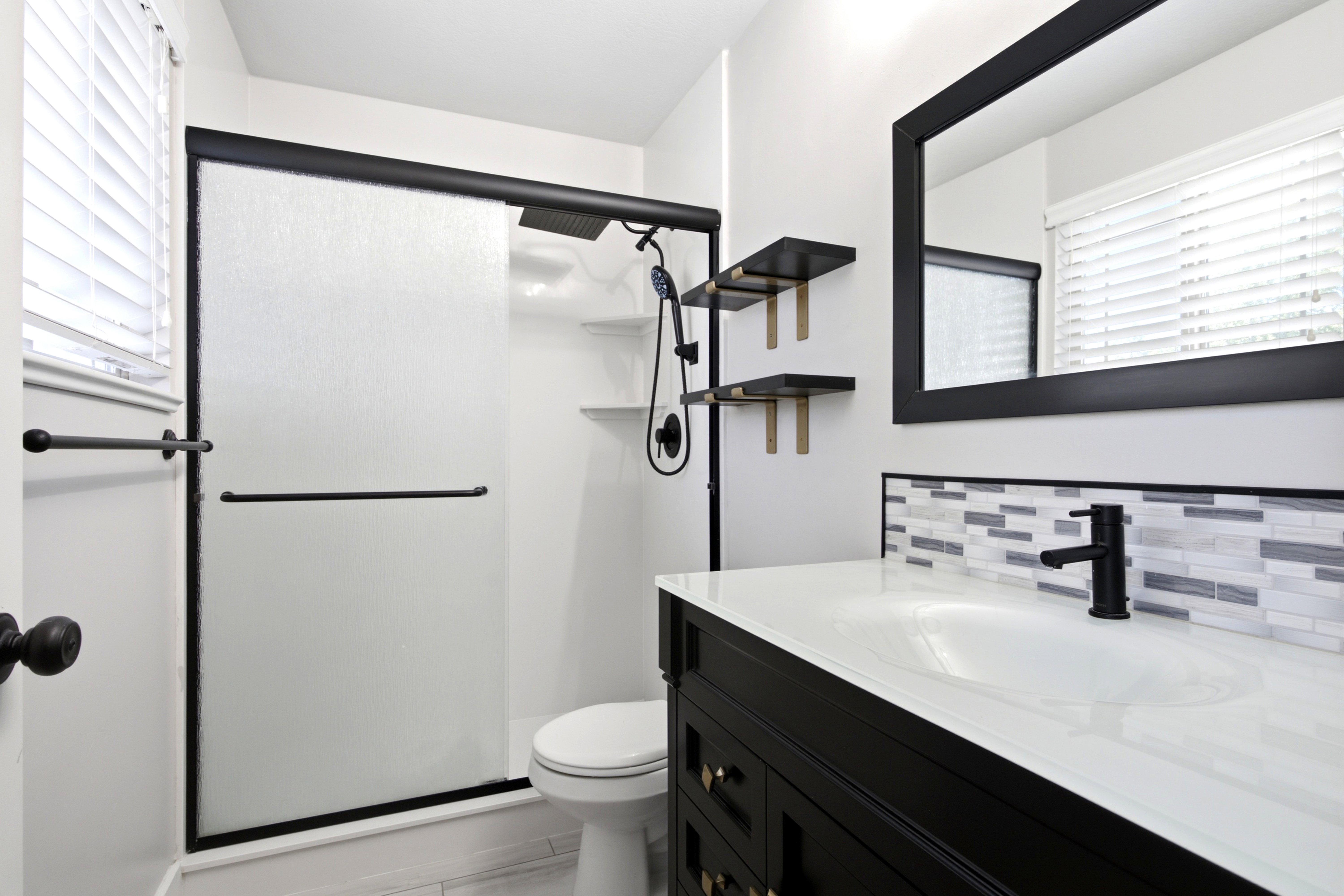 Contemporary bathroom featuring a dark vanity with a white countertop, mosaic tile backsplash, a large framed mirror, and a shower with a sliding frosted glass door