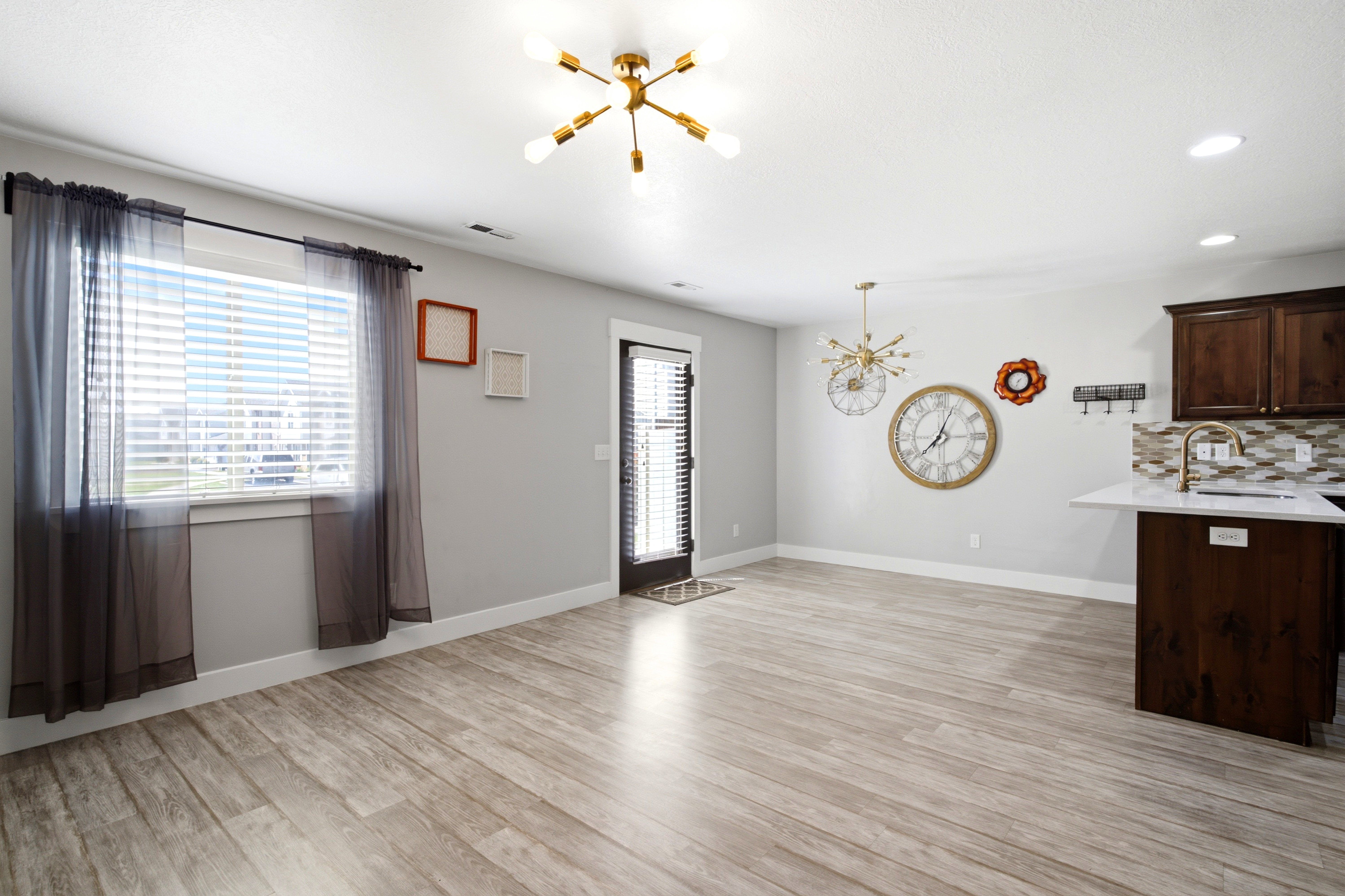 Open-concept living space featuring wood-finish flooring, a modern sputnik light fixture, a separate recessed light, a window with blinds, and a kitchen peninsula with dark wood cabinetry and a mosaic tile backsplash