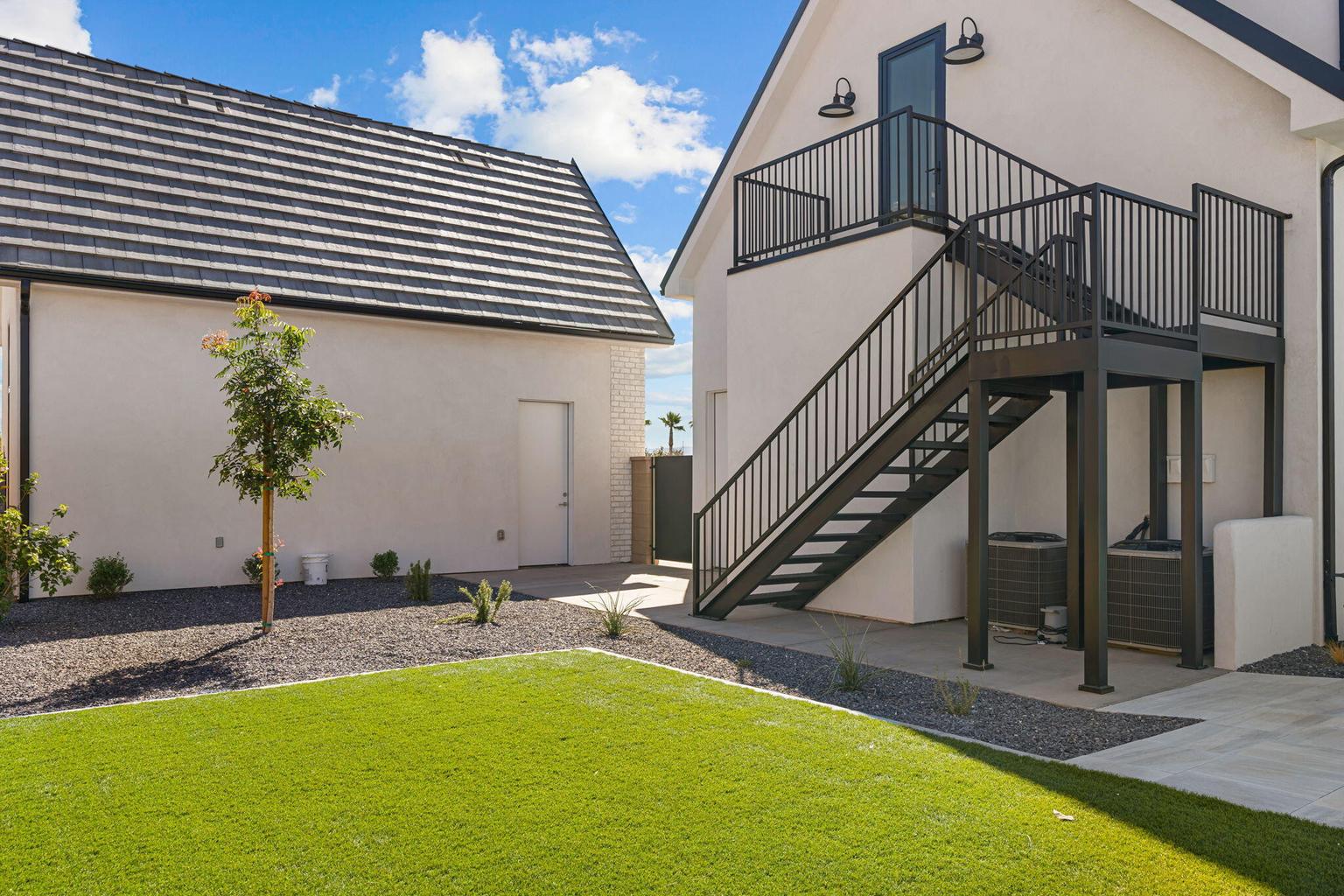 Exterior featuring a modern black metal staircase providing access to an upper-level balcony, a stucco finish facade, a dark tile roof, and a manicured lawn with surrounding gravel landscaping