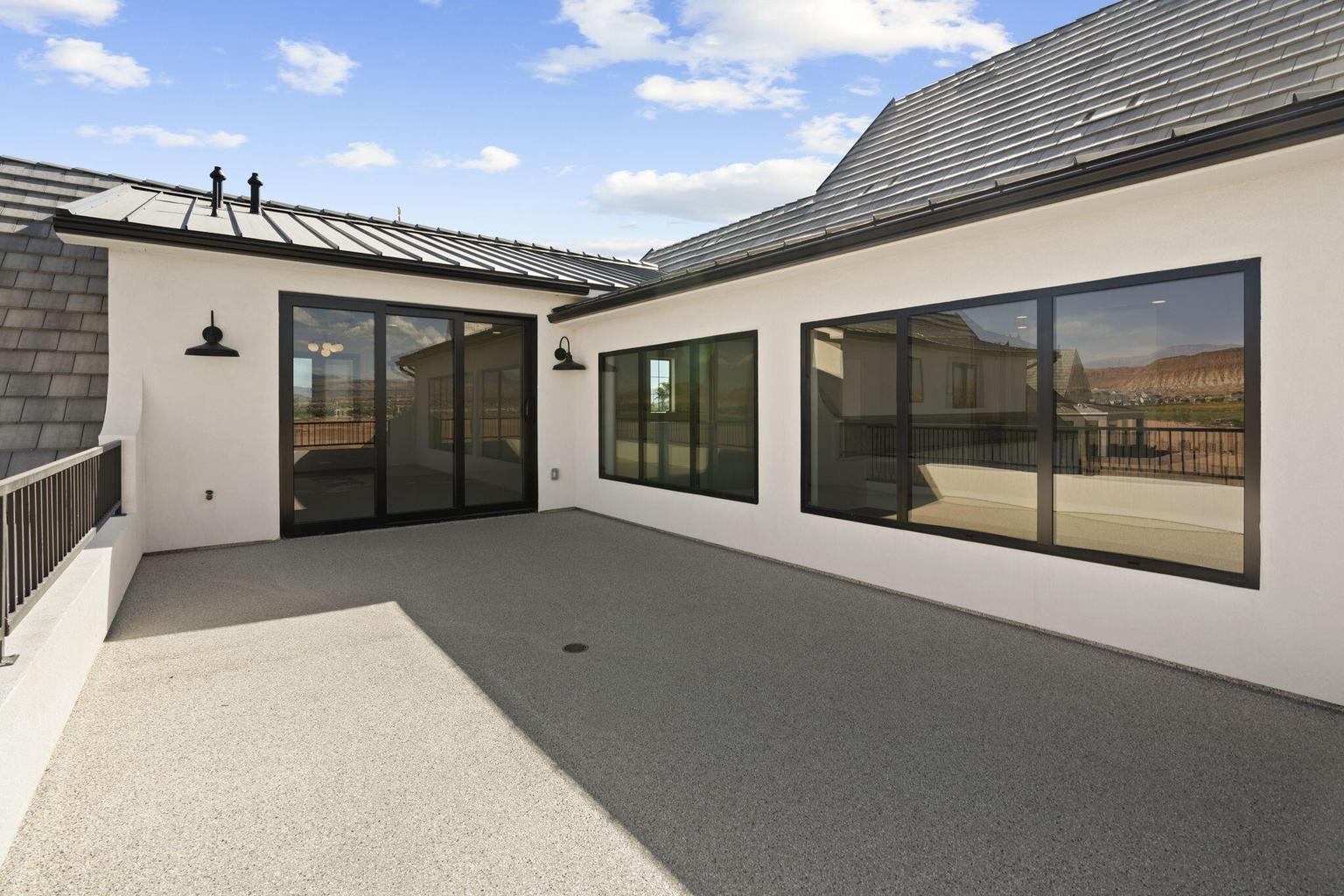 Expansive upper-level patio featuring a textured concrete surface, modern black-framed sliding glass doors, large picture windows, and exterior sconce lighting