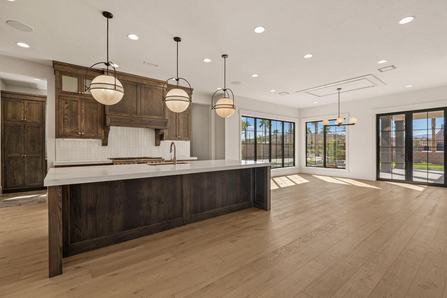 Open-concept kitchen and living area featuring a large center island with a light-toned countertop, rich wood-finish cabinetry, a white subway tile backsplash, light wood-finish flooring, and multiple large windows with black frames