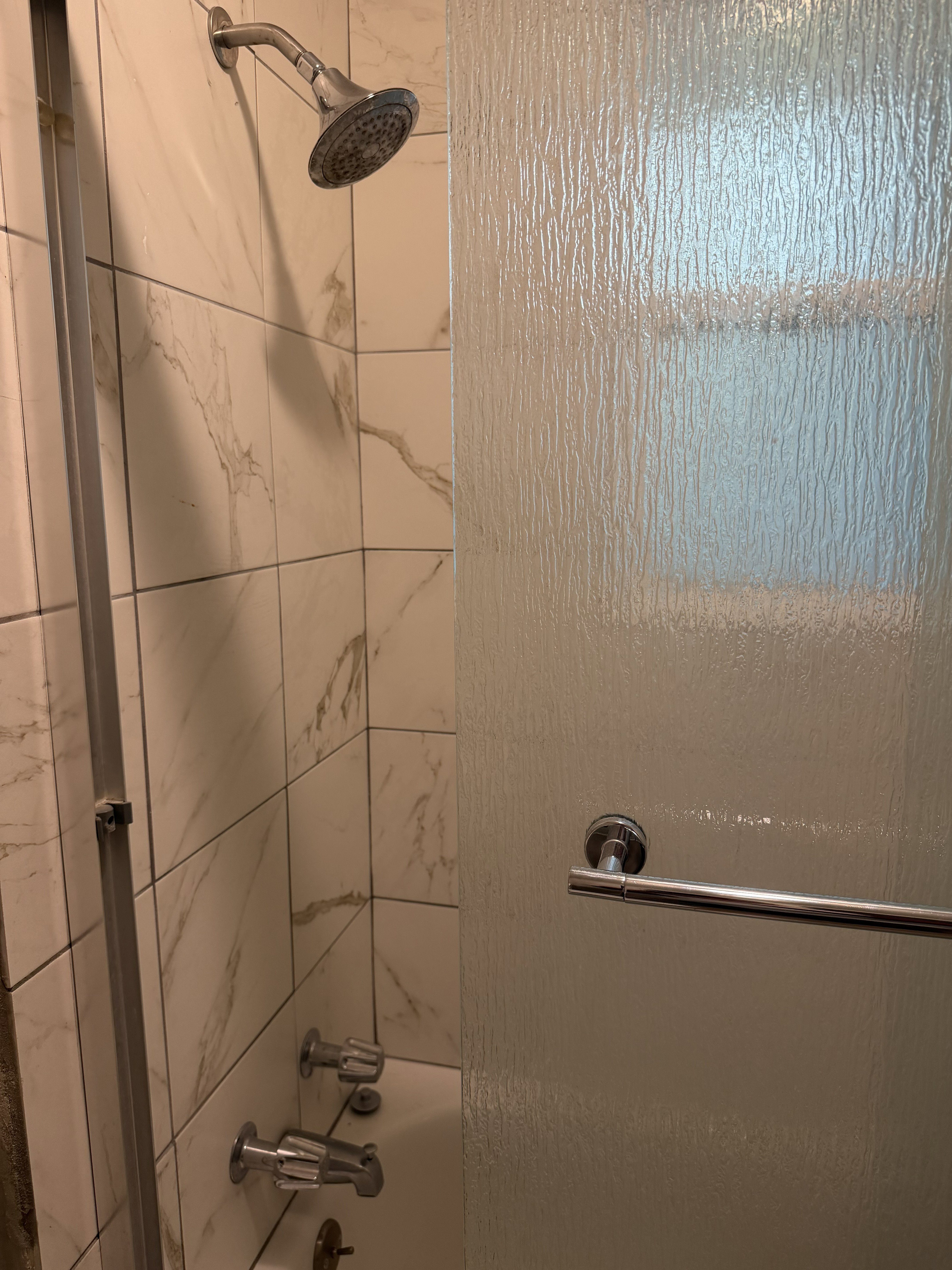Bathtub and shower combination featuring white tiled walls with subtle veining, a chrome showerhead, and a textured privacy glass enclosure with a chrome towel bar