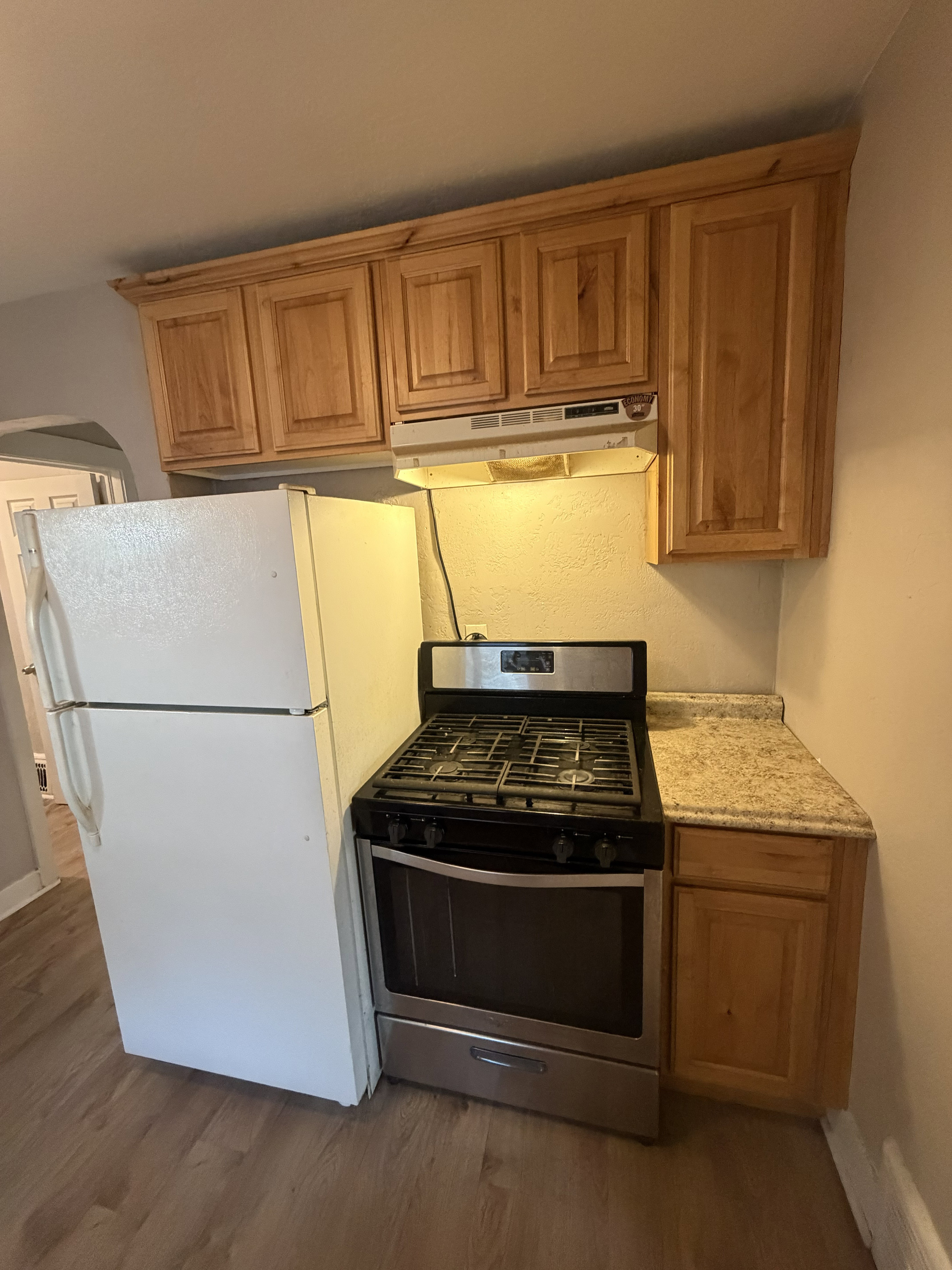 Kitchenette featuring wood-finish cabinetry with crown molding, a stainless steel gas range, a coordinating range hood, and a light-toned speckled countertop