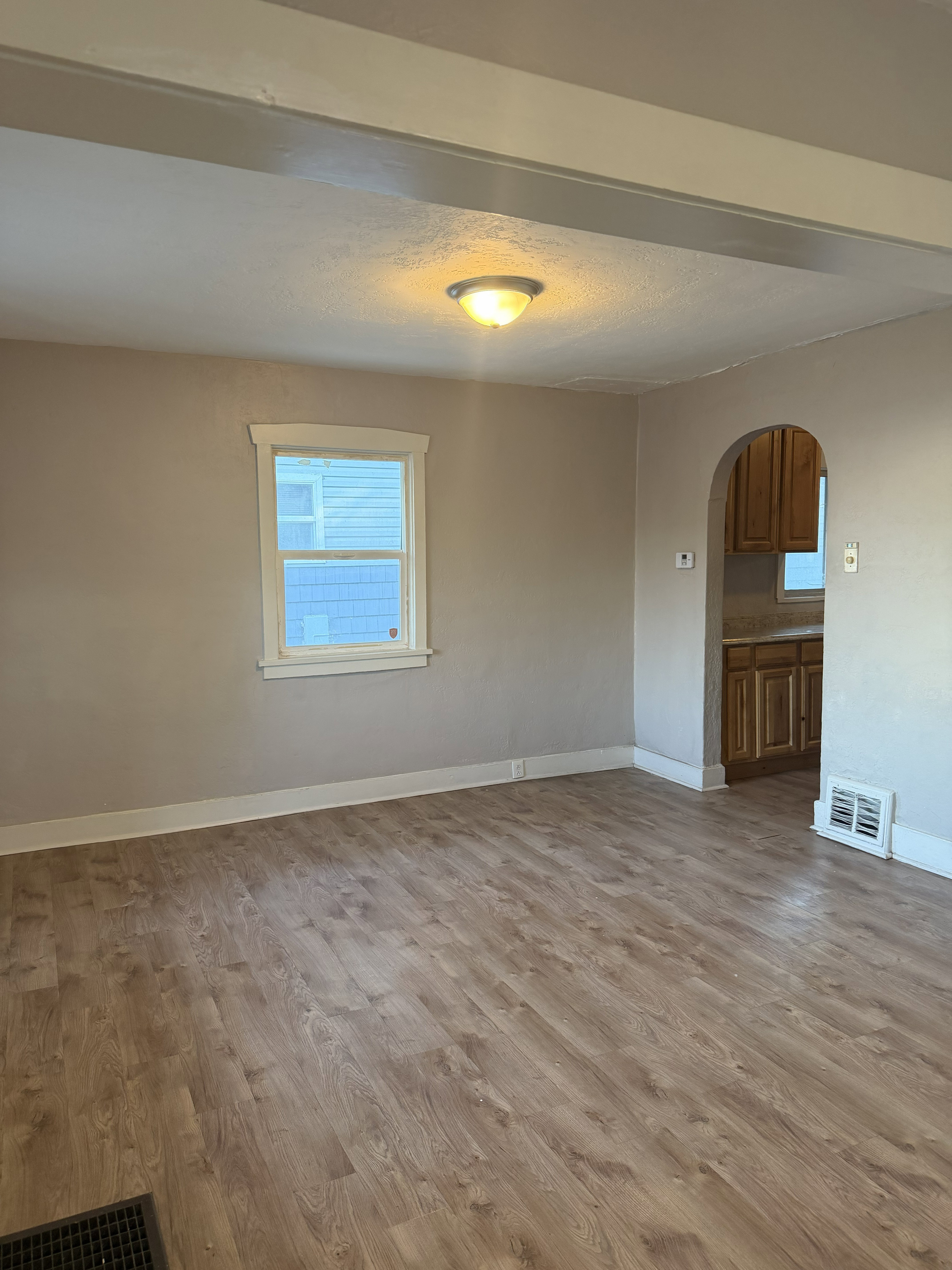 Spacious room featuring wood-finish flooring, a ceiling-mounted light fixture, a window with white trim, a floor vent, and an arched entryway to an adjacent space with wood cabinetry