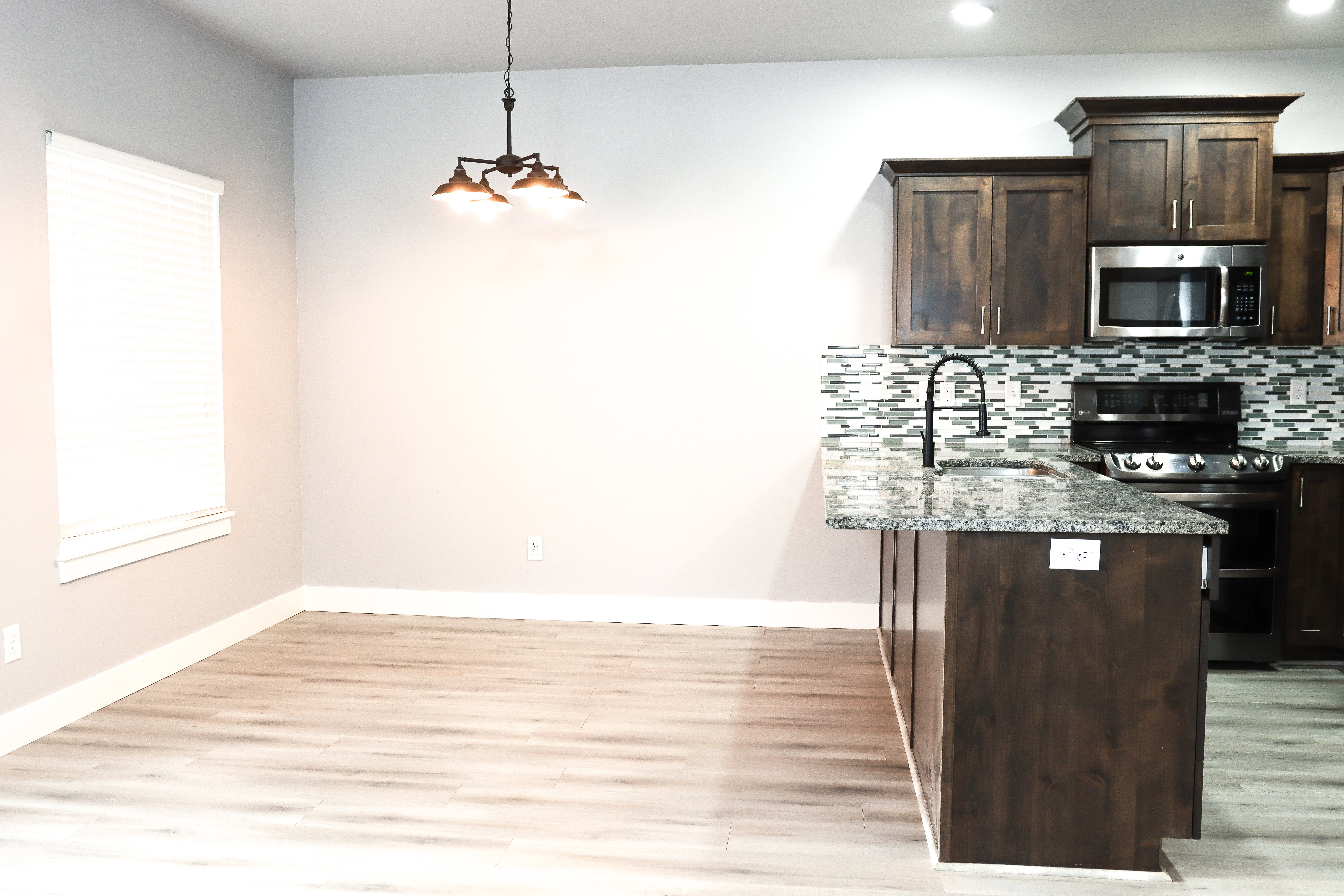 Open concept kitchen and dining area featuring wood-finish flooring, dark wood cabinetry, stainless steel appliances, a tile backsplash, and an island with a built-in sink