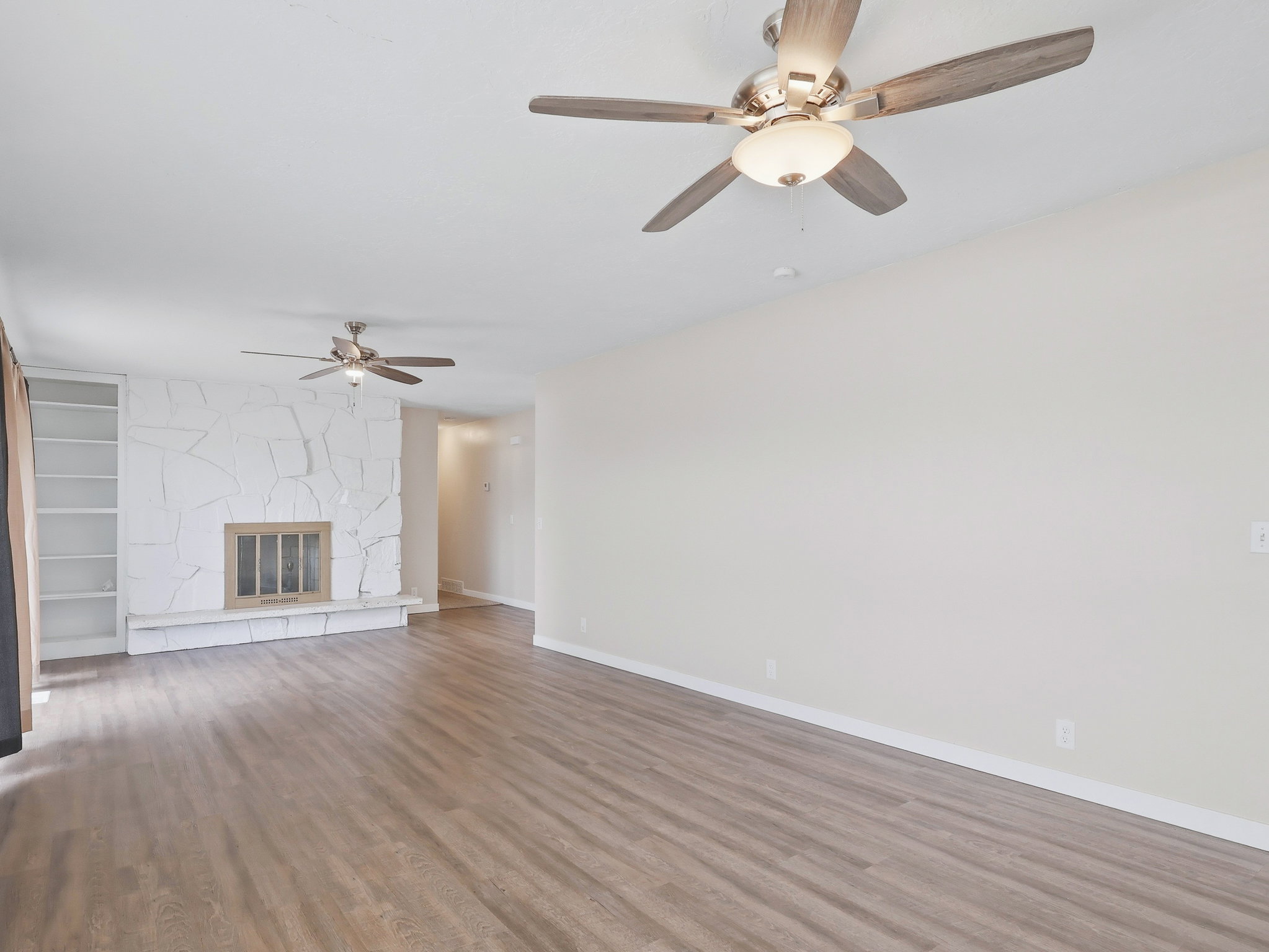 Open-concept living area featuring wood-finish flooring, a white stone fireplace with a hearth, built-in shelving, and two ceiling fans with integrated lighting
