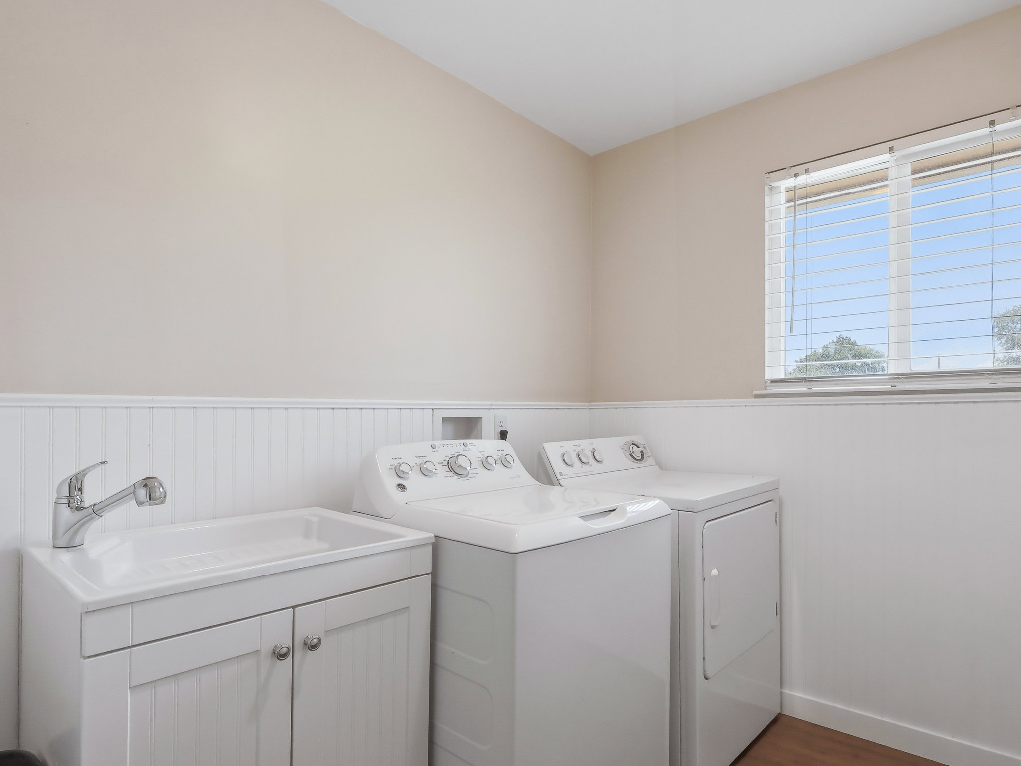 Dedicated laundry area featuring a utility sink, white base cabinetry with brushed nickel hardware, beadboard wainscoting, and a window with blinds