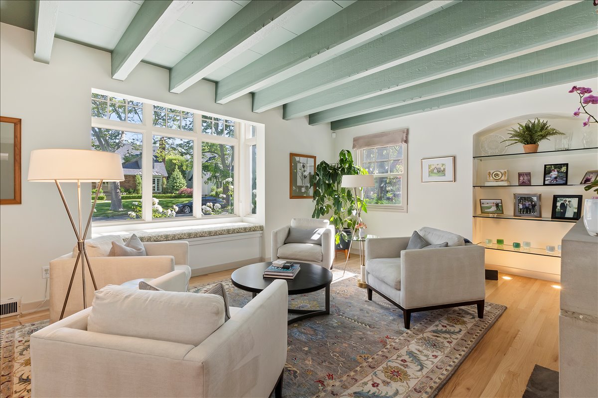 Living area featuring a large picture window with a built-in window seat, exposed ceiling beams, hardwood flooring, and an arched alcove with glass shelving and integrated lighting
