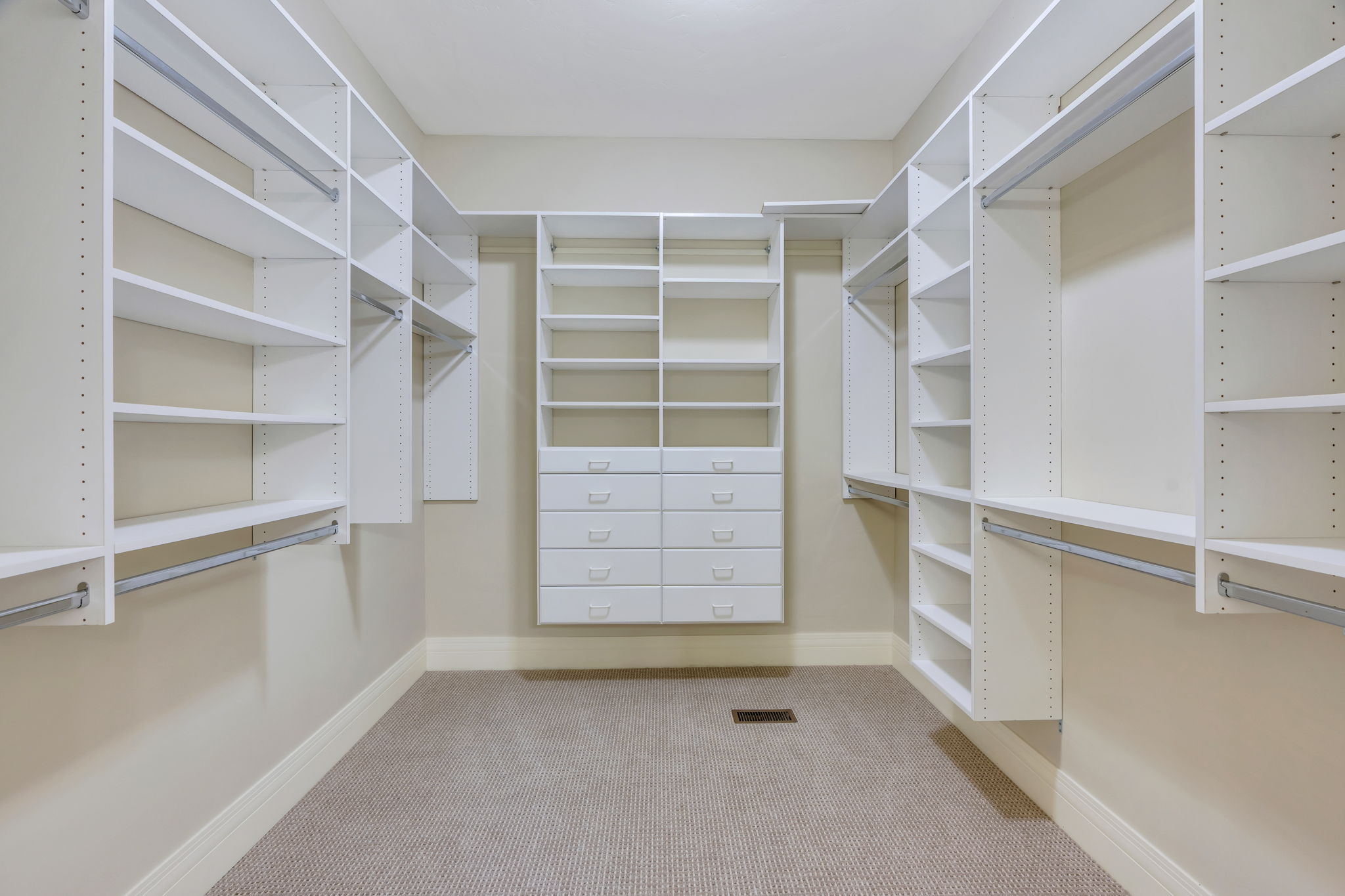Primary bedroom walk-in closet with extensive white built-in shelving, multiple hanging rods, and a central dresser unit featuring multiple drawers