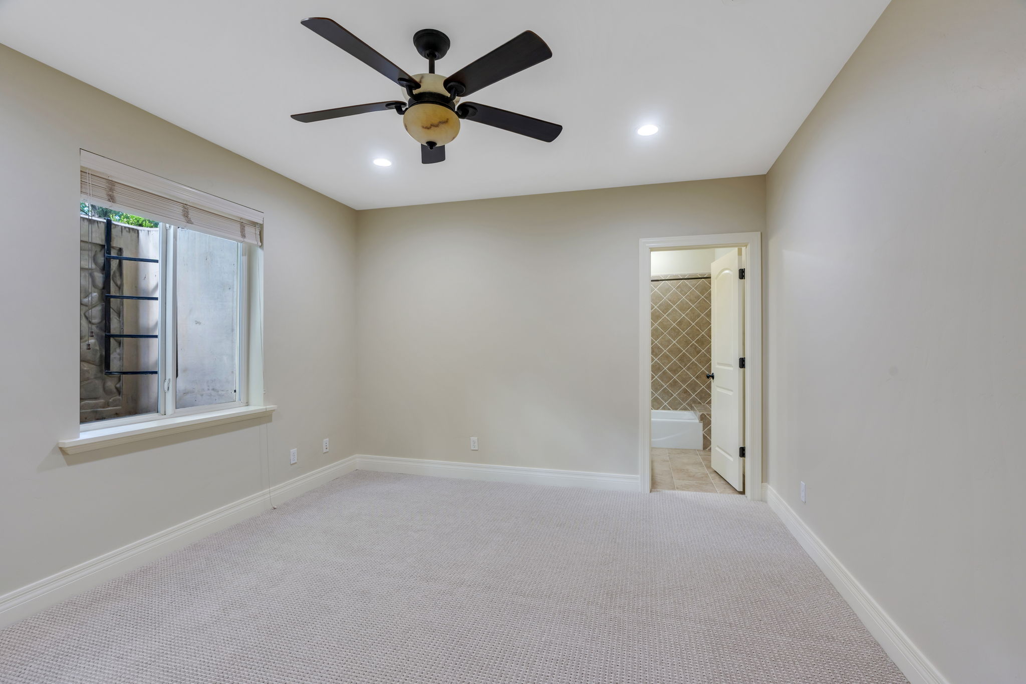 One of two bedrooms in basement featuring  ceiling fans with integrated lighting, recessed ceiling lights, a window with a stone-lined window well, and an ensuite bathroom with tiled shower and flooring