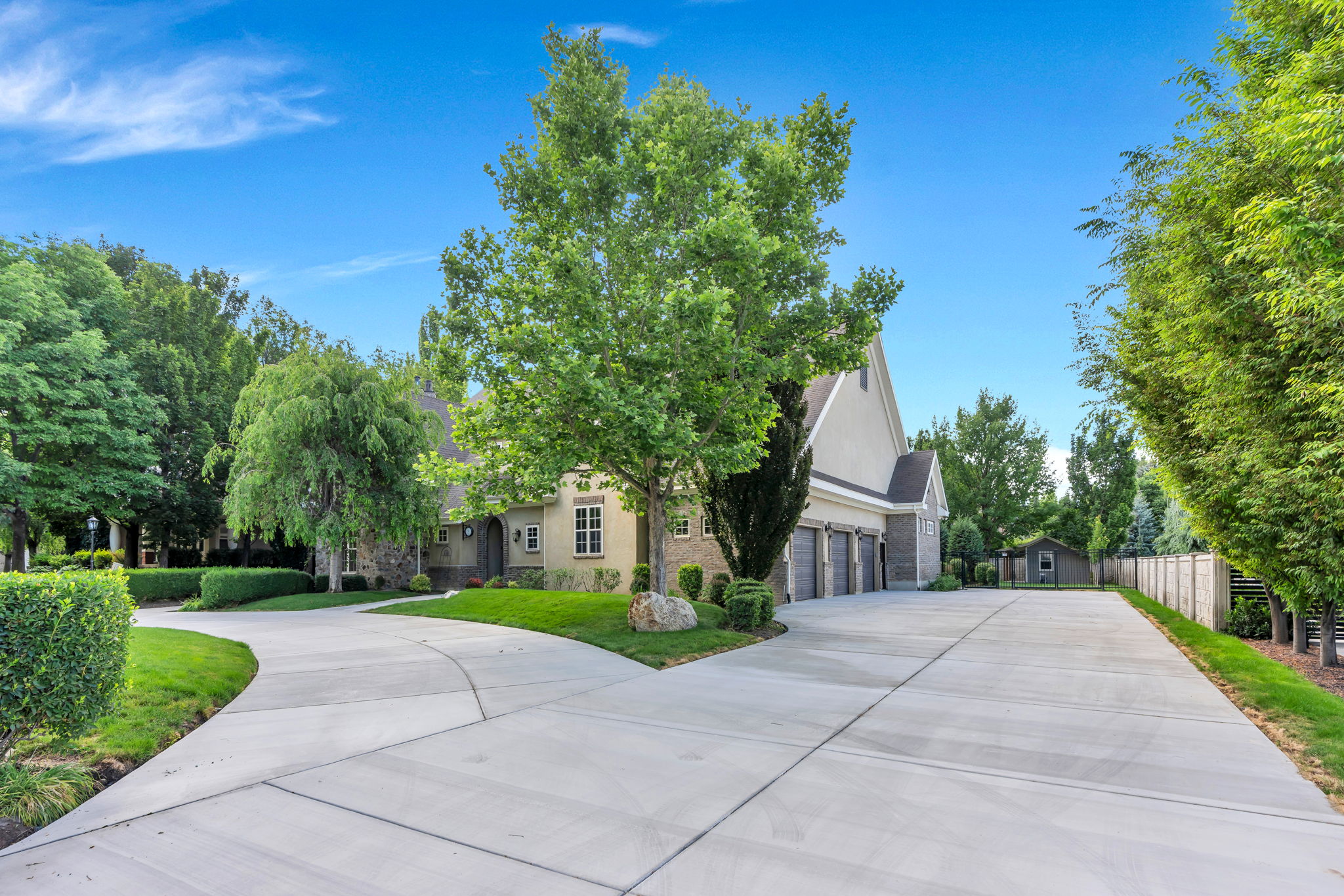 Expansive concrete driveway with a circular drive, lush green landscaping, mature trees, and a multi-car garage