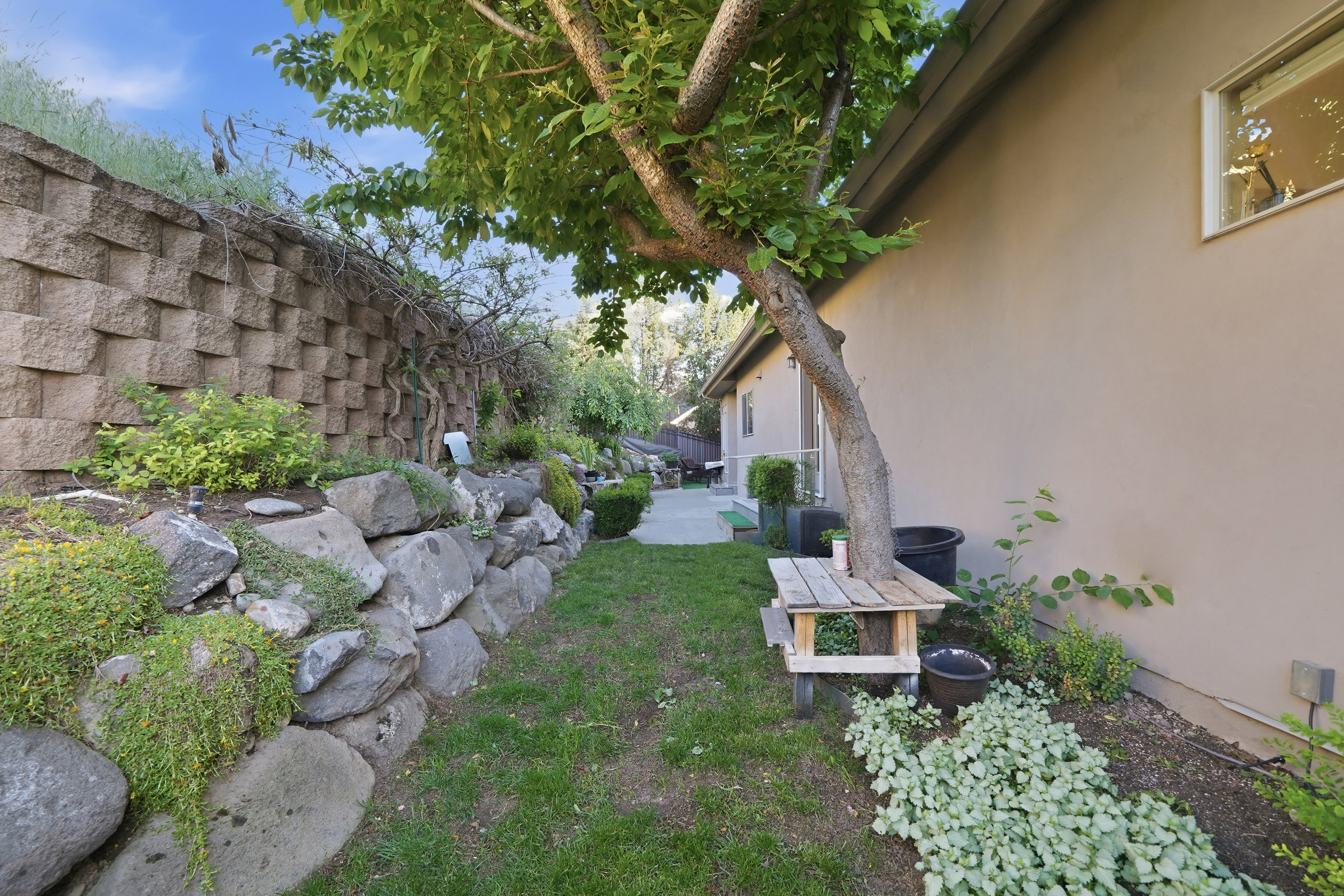 Lush side yard featuring a decorative retaining wall, natural rock landscaping, mature tree, manicured lawn, and stucco exterior