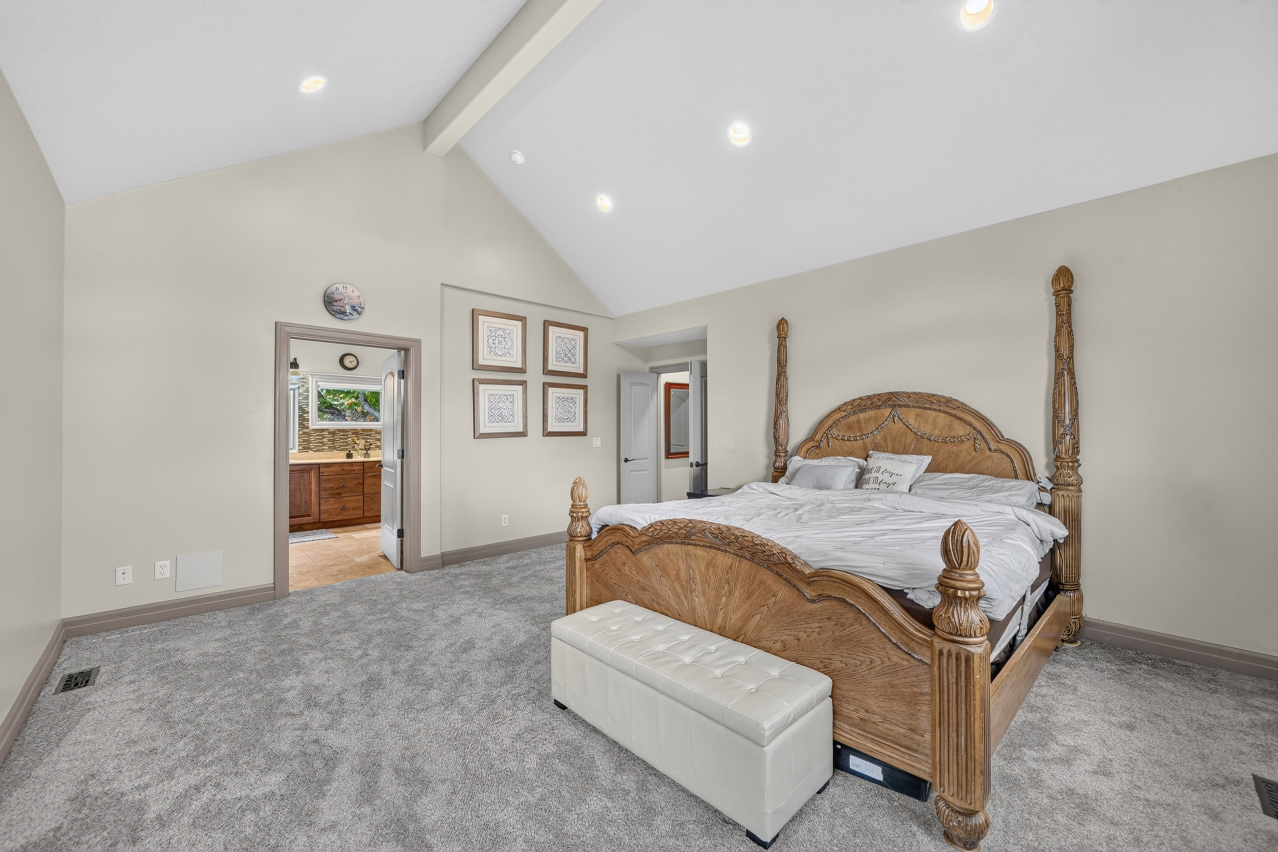 Vaulted ceiling bedroom with recessed lighting, light gray walls, gray carpet flooring, and a doorway revealing a bathroom with a wood vanity and window