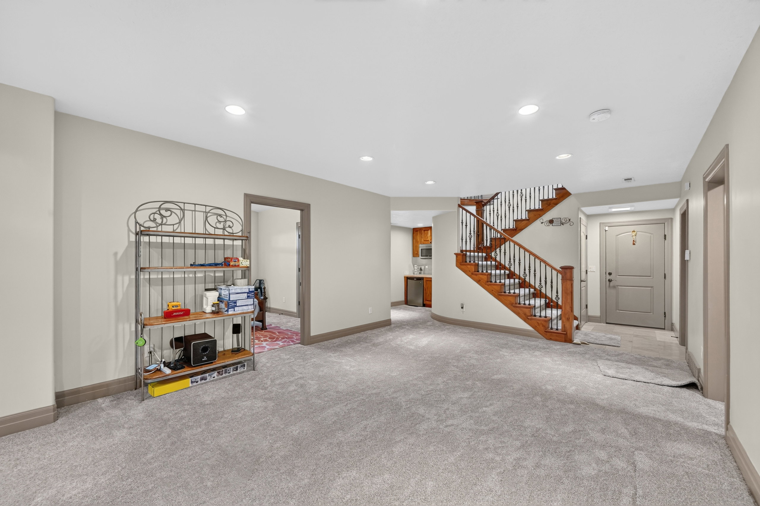 Spacious carpeted area with recessed lighting, an open wood staircase with wrought iron balusters, a built-in wet bar with wood cabinetry and a stainless steel sink, and a solid panel door