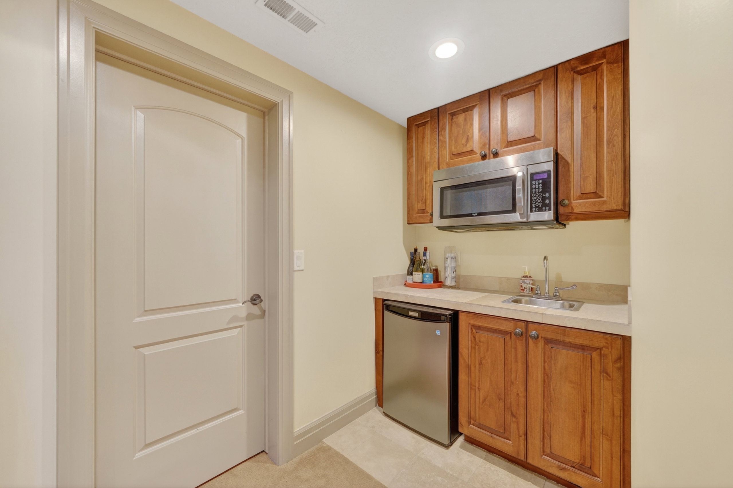 Compact wet bar featuring wood cabinetry, a stainless steel microwave, a mini-refrigerator, a sink, and a light-toned countertop