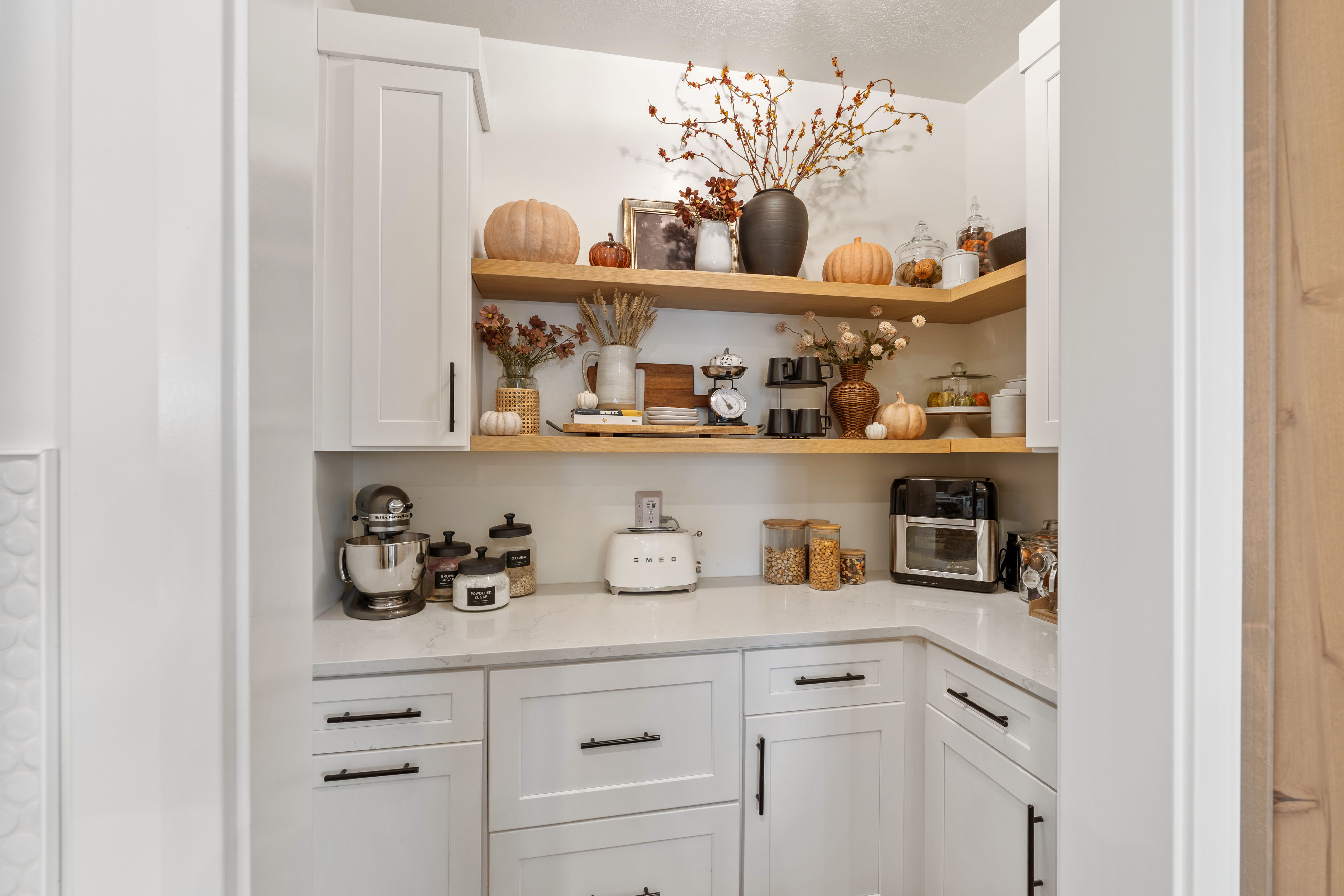Gorgeous corner pantry featuring white cabinetry with black hardware, open wood shelving, a light-toned countertop, and white walls