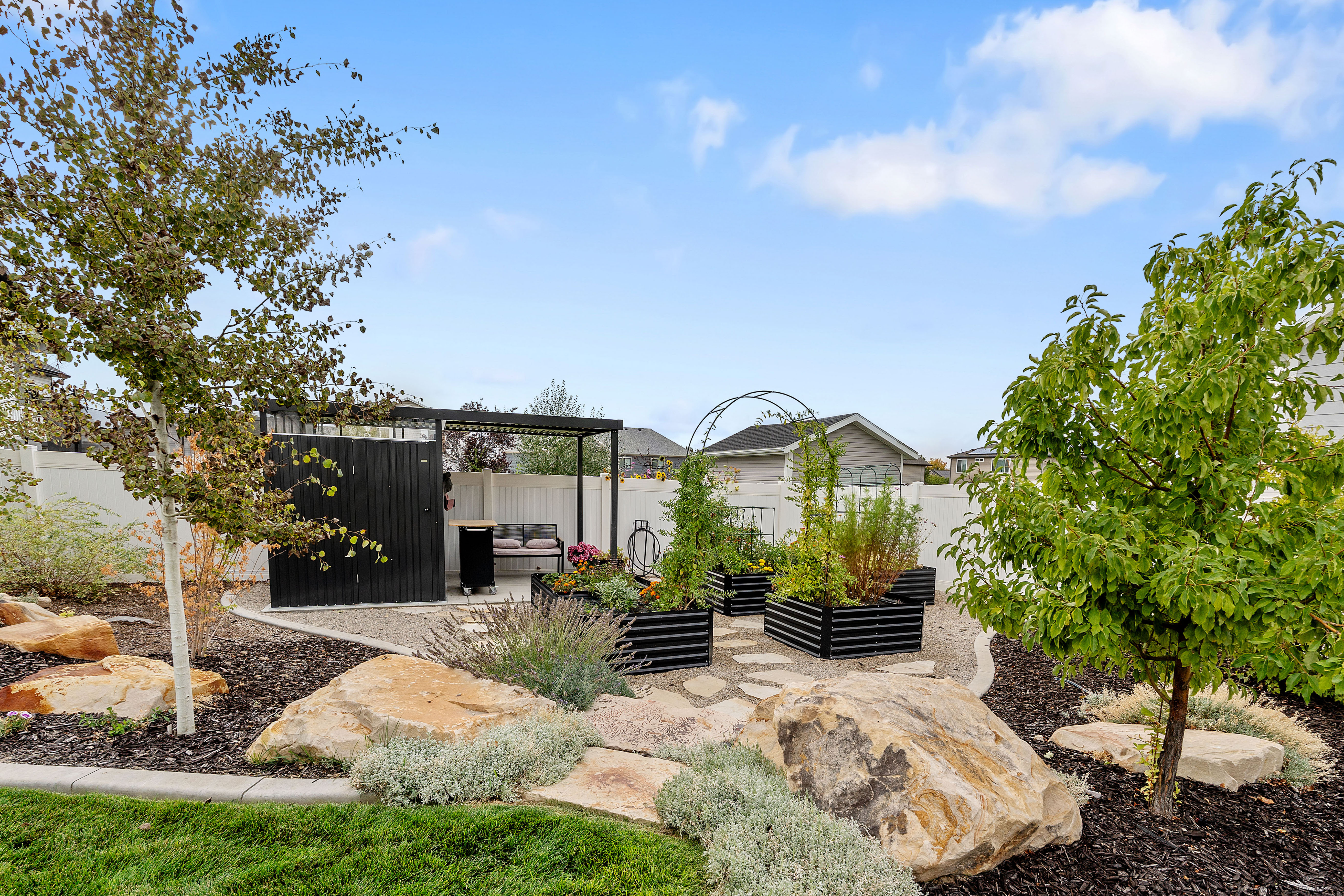 Landscaped backyard featuring decorative rockery, mature trees, and a gravel patio with a black privacy screen and pergola
