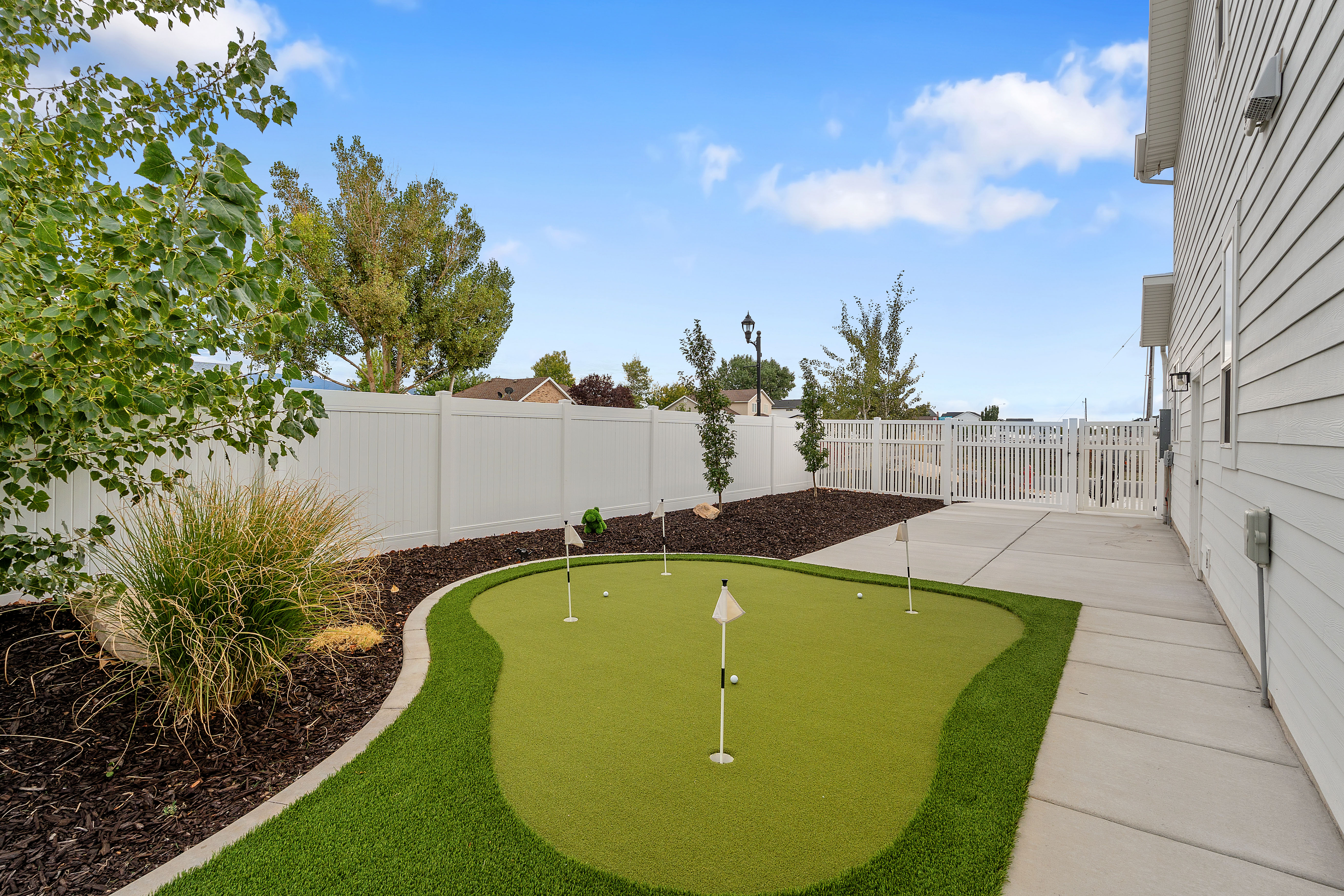 Backyard putting green with artificial turf, bordered by a concrete pathway and a white privacy fence