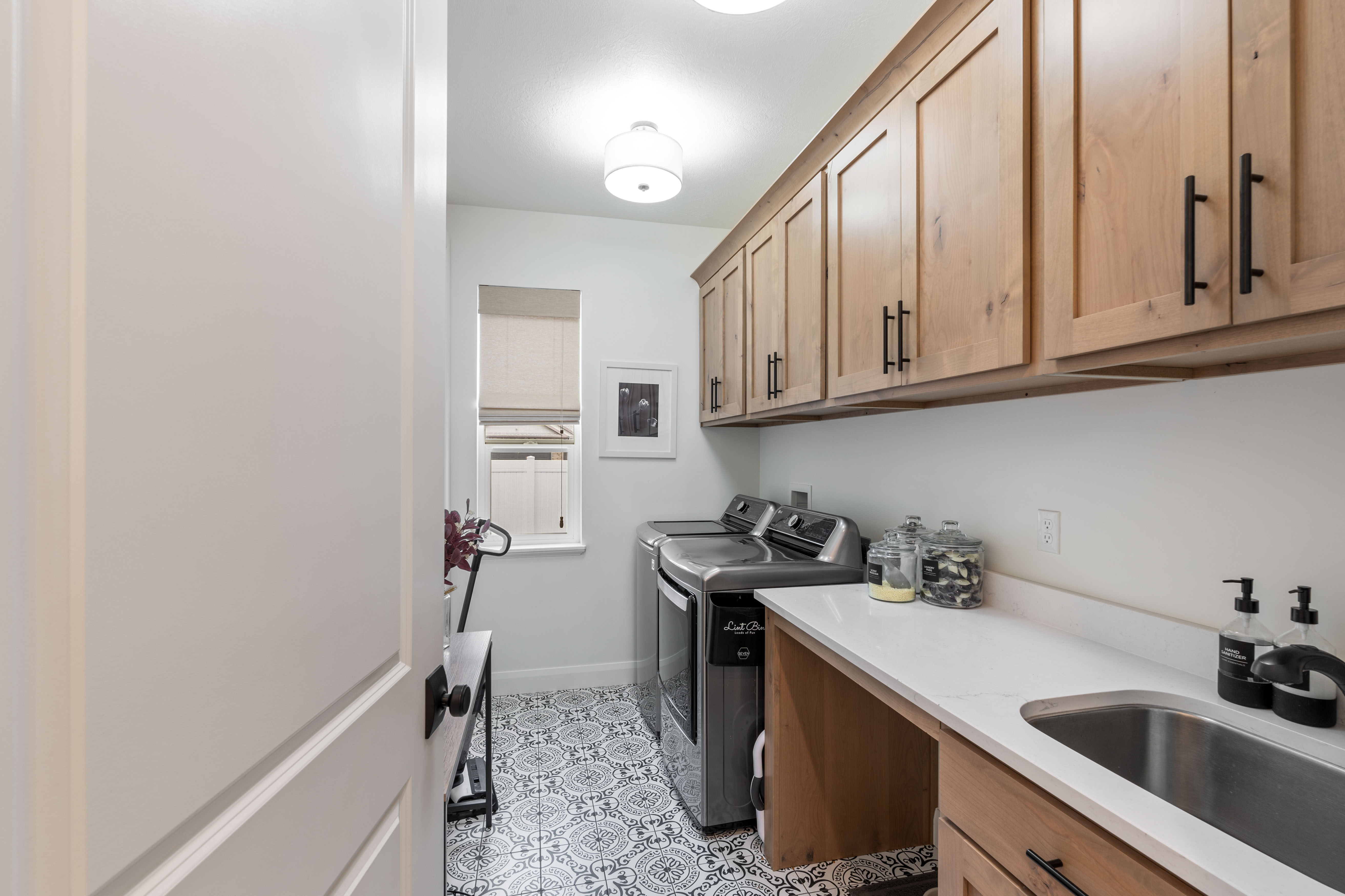 Laundry room featuring patterned floor tiling, natural wood cabinetry with matte black hardware, a built-in stainless steel sink, light-colored countertops, and a window for natural illumination