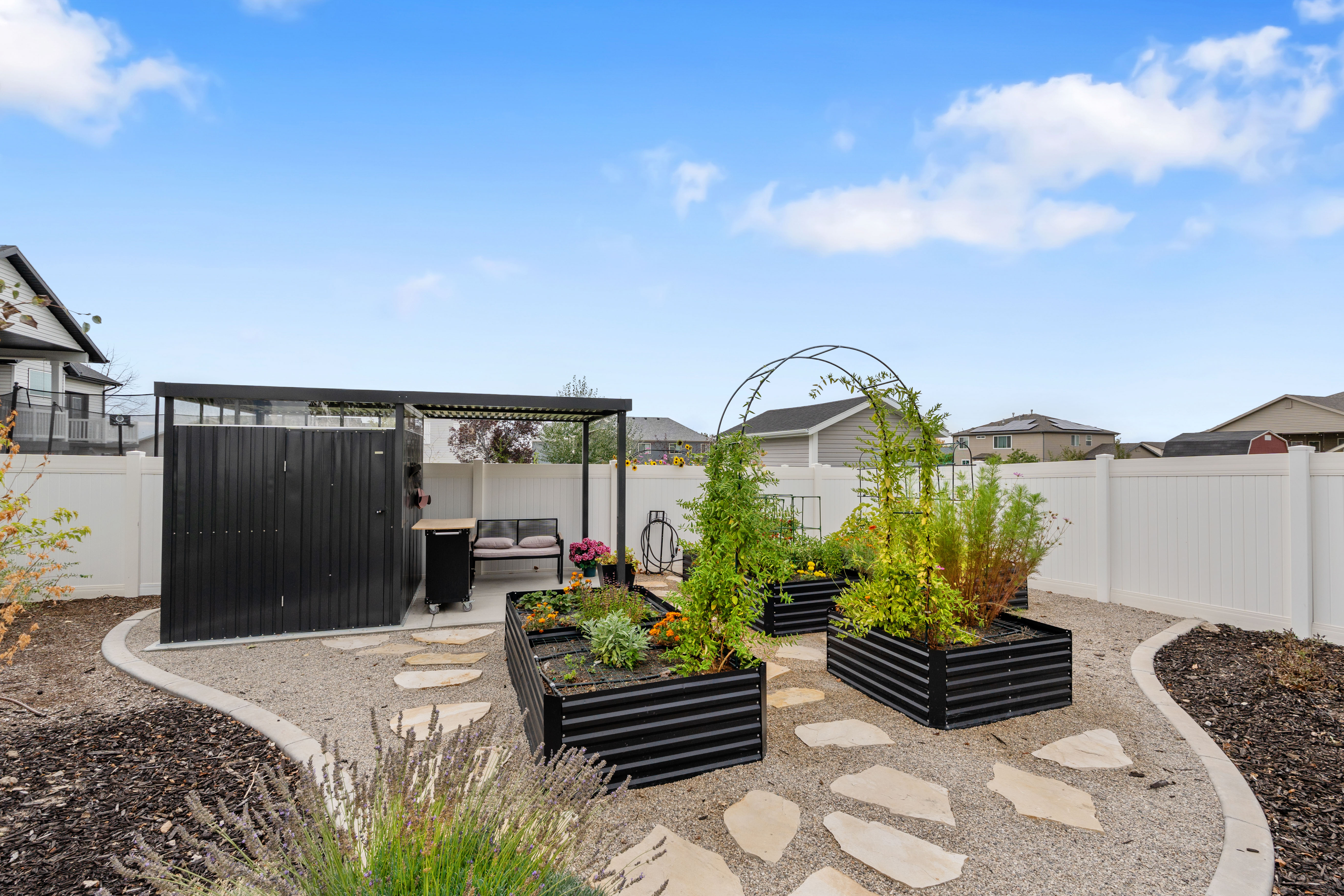 Garden shed and raised beds - Expansive backyard featuring a white vinyl privacy fence, a covered patio area with a pergola, a dedicated gardening space with raised planters, a gravel pathway with stepping stones, and dark mulch landscaping
