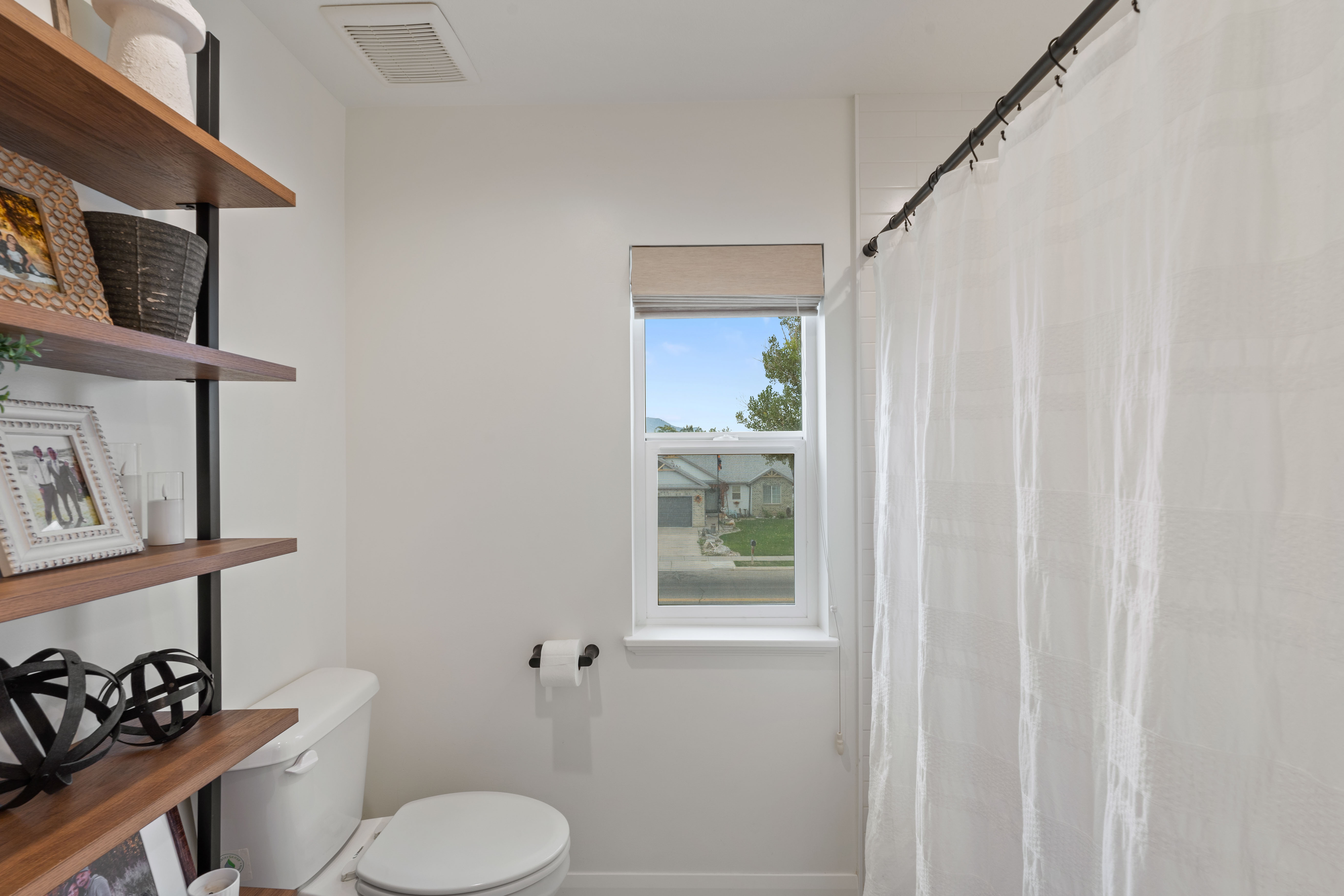 Bathroom featuring an elongated toilet, a single pane window with a roller shade, and wall-mounted shelving with wood-finish shelves