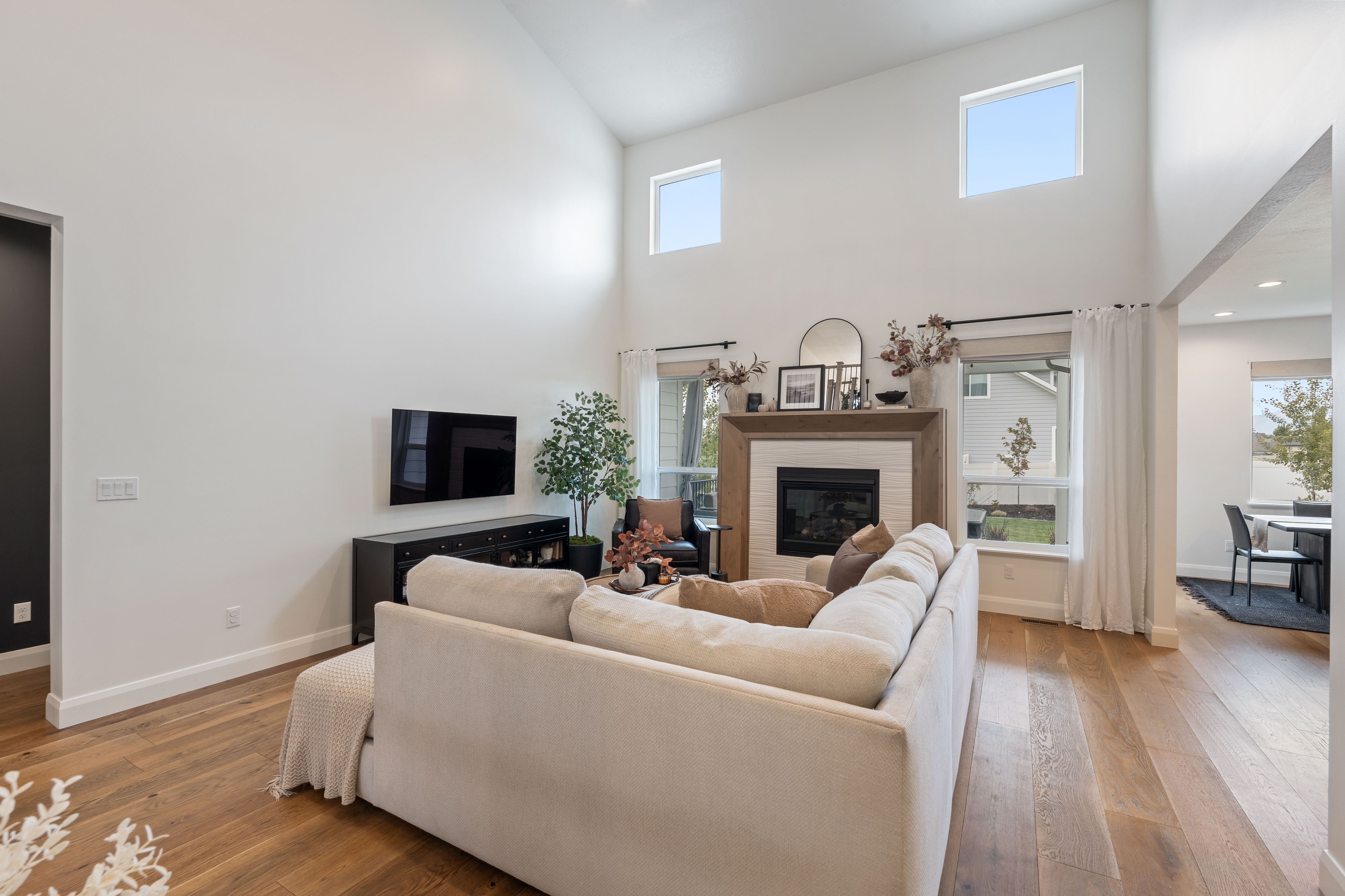 Soaring ceilings with upper windows, central fireplace with wood surround, wide-plank wood-finish flooring, recessed lighting, and large windows with privacy blinds