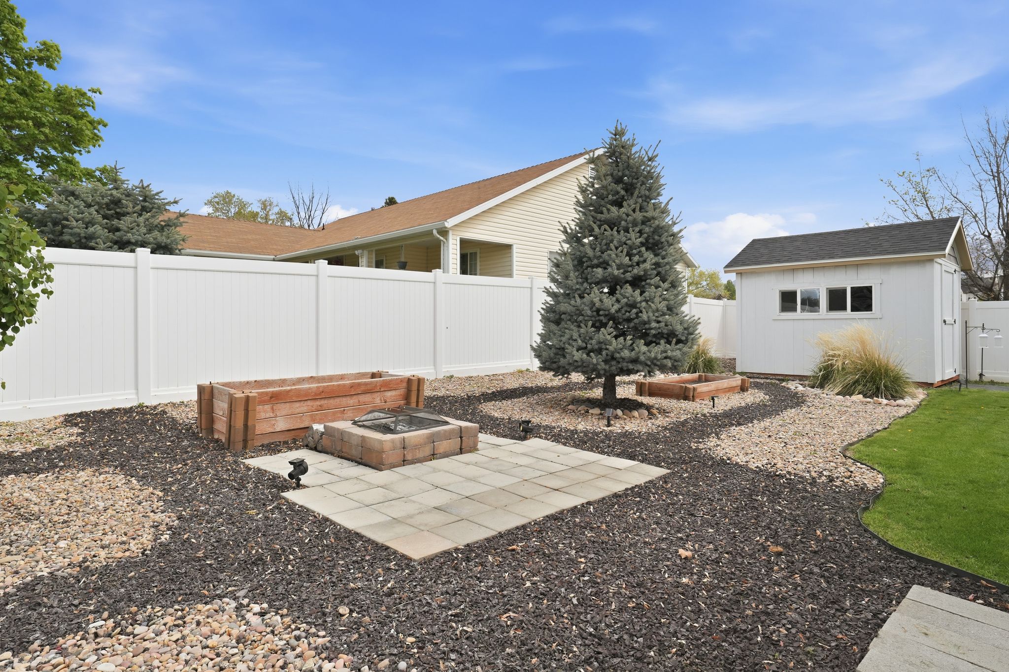 Expansive backyard featuring a white vinyl privacy fence, a detached storage shed with multiple windows, a mature evergreen tree, and a paved patio area with a built-in fire pit
