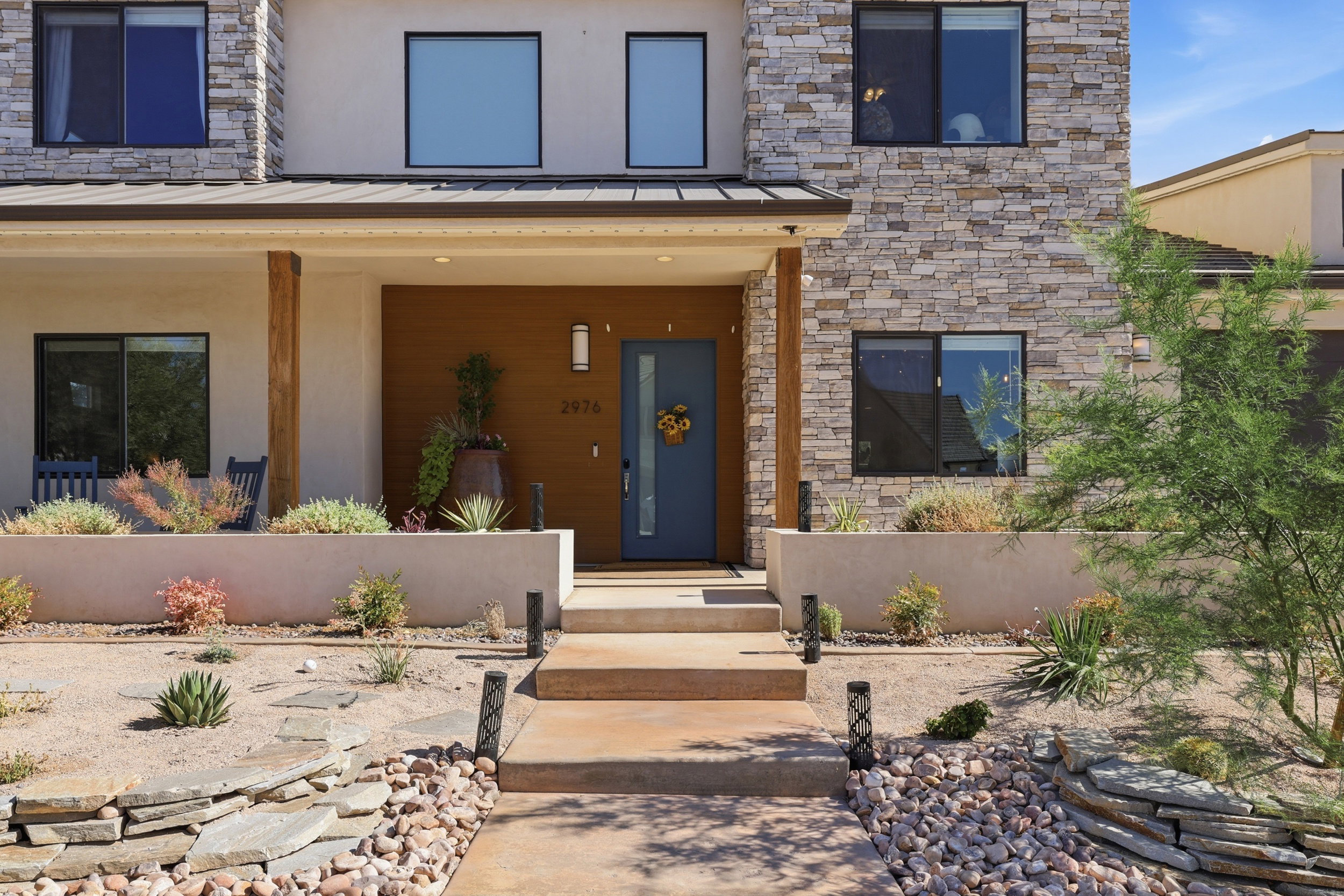 Modern facade featuring a blue entry door, stone and stucco exterior, wood-paneled accents, and desert-inspired landscaping with stone pathways and planters