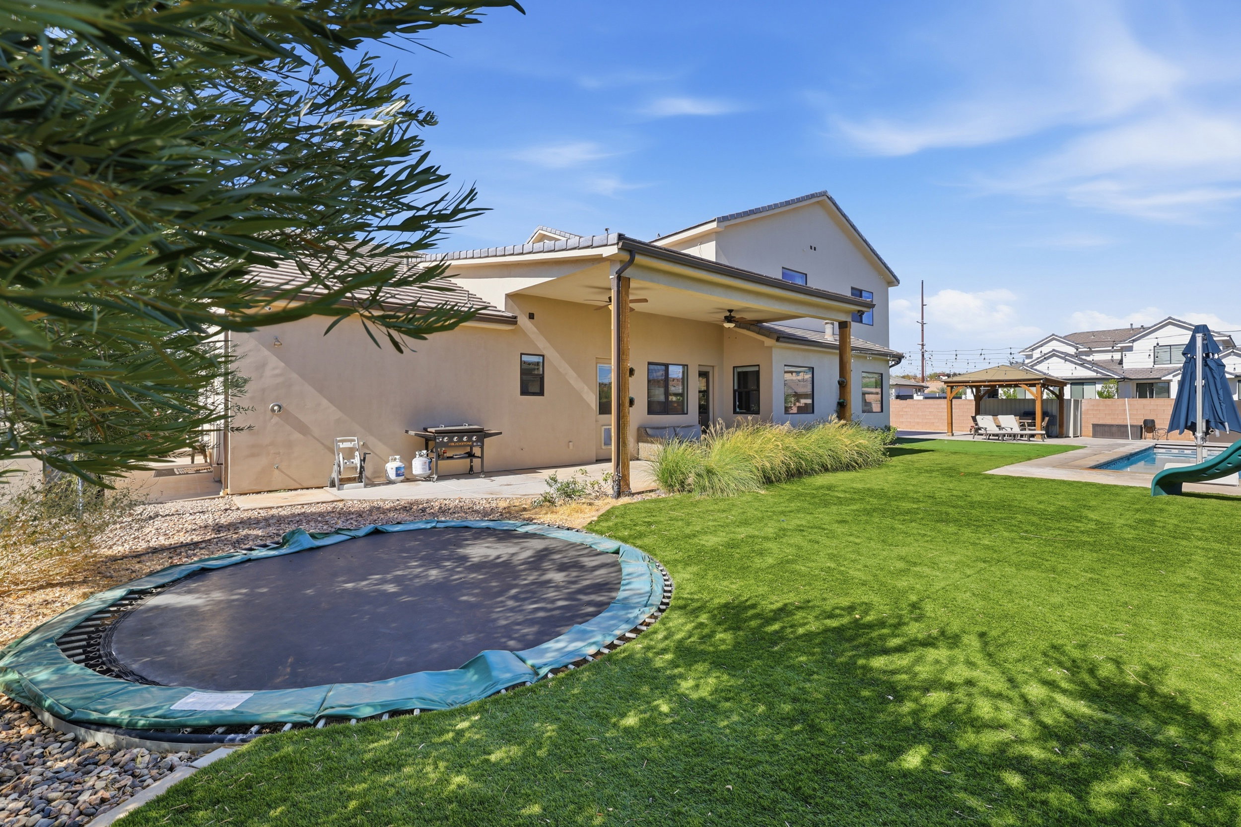 Expansive backyard featuring artificial turf, a covered patio with columns, a swimming pool with a slide, an adjacent pergola, and a built-in trampoline