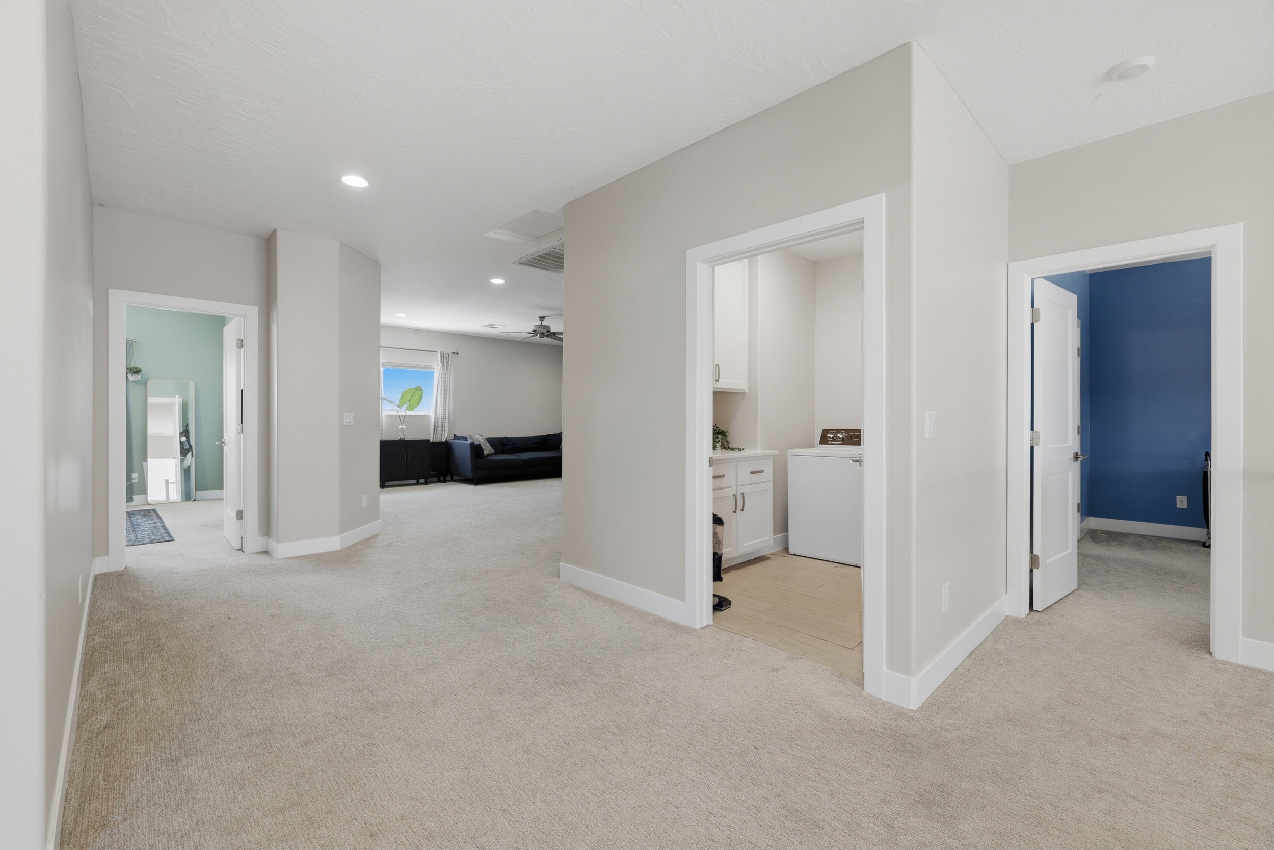 Spacious carpeted hallway with recessed lighting and white baseboards, offering access to a laundry area with white cabinetry, a room with a blue accent wall, and another room with light blue walls