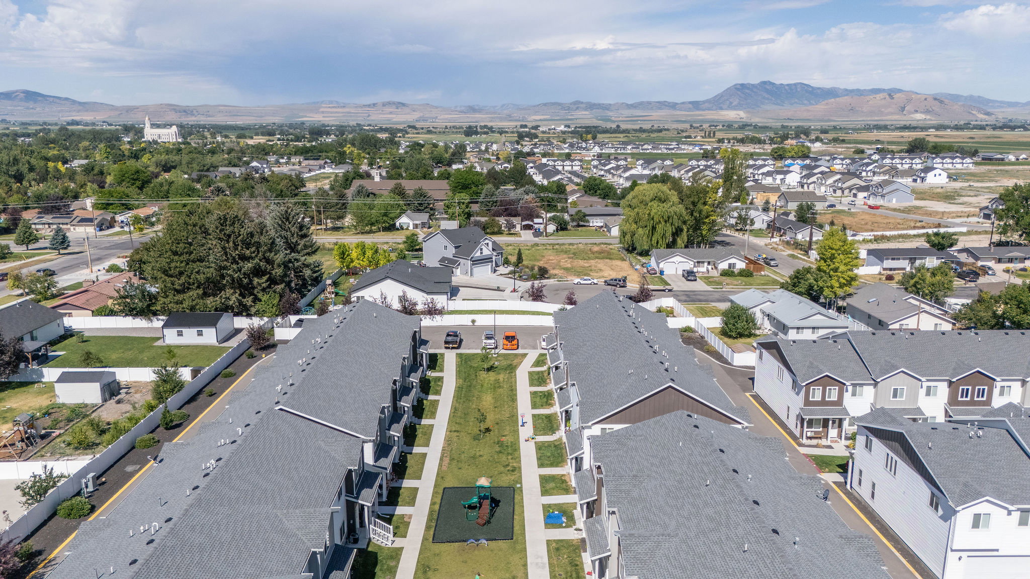 Aerial perspective showcasing a community of townhomes with gray shingle roofs and private patios, featuring a central green space with a playground