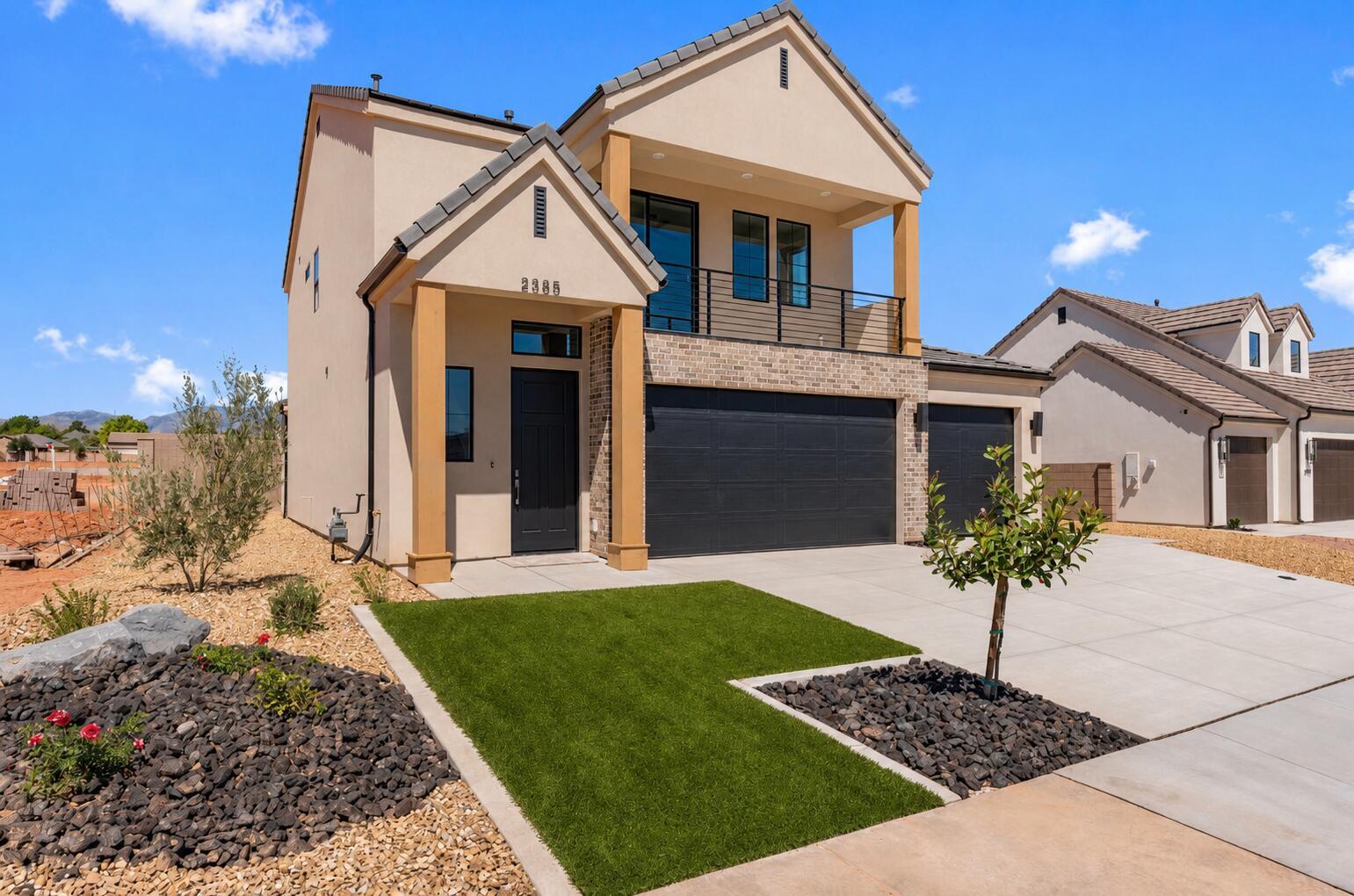 Contemporary stucco exterior featuring a stone accent wall, two-car garage, covered entry, second-story balcony with metal railing, and low-maintenance landscaping with artificial turf and decorative rock