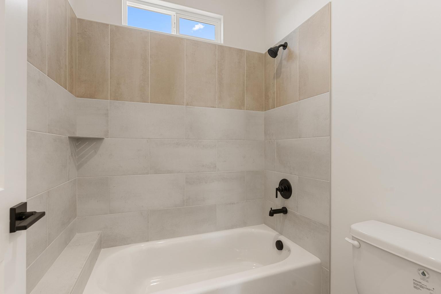 Bathtub with matte black fixtures, two-tone tiled shower surround, recessed shower shelf, and a high transom window