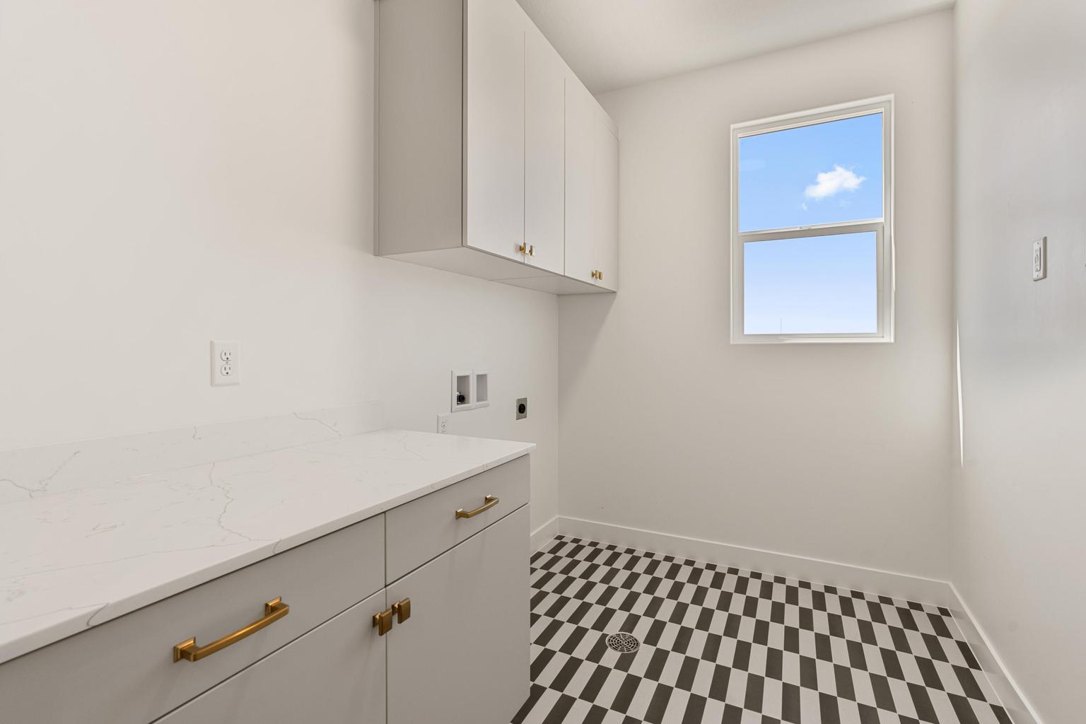 Laundry area featuring a large window, contrasting geometric floor tile, white shaker-style cabinetry, light-toned countertop, and brass hardware