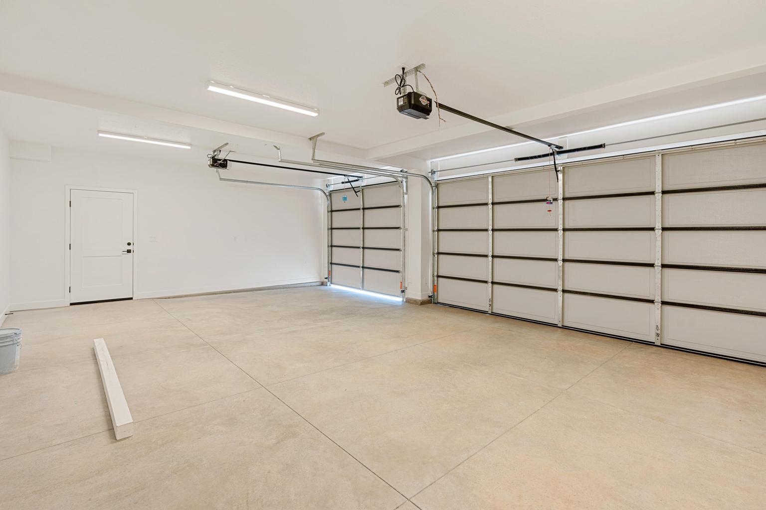 Spacious garage featuring two sectional garage doors with automatic openers, a side entry door, concrete flooring, and overhead strip lighting