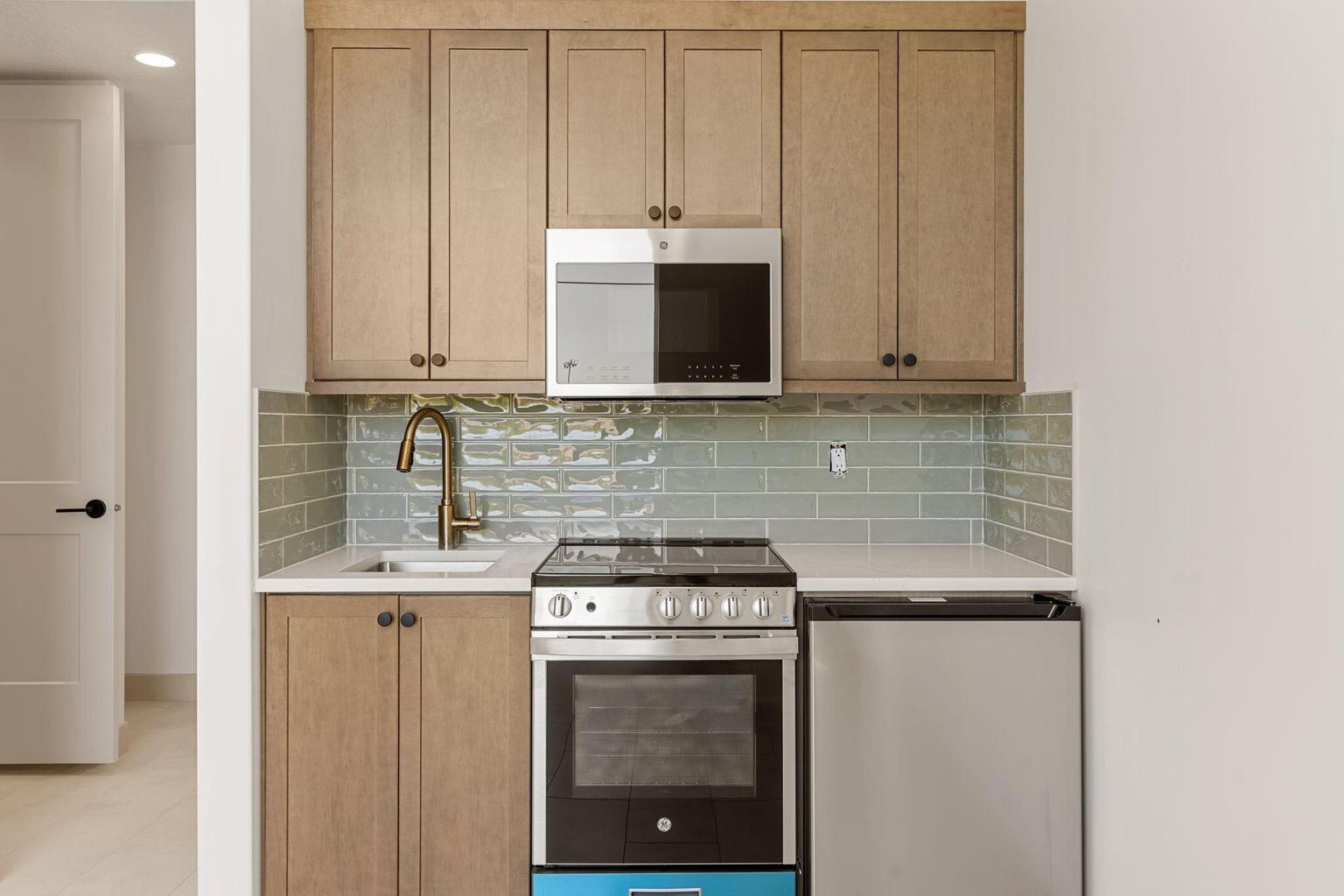 Compact kitchen space featuring light wood-finish cabinetry, a stainless steel microwave, a built-in electric range, a stainless steel refrigerator, and a light blue subway tile backsplash