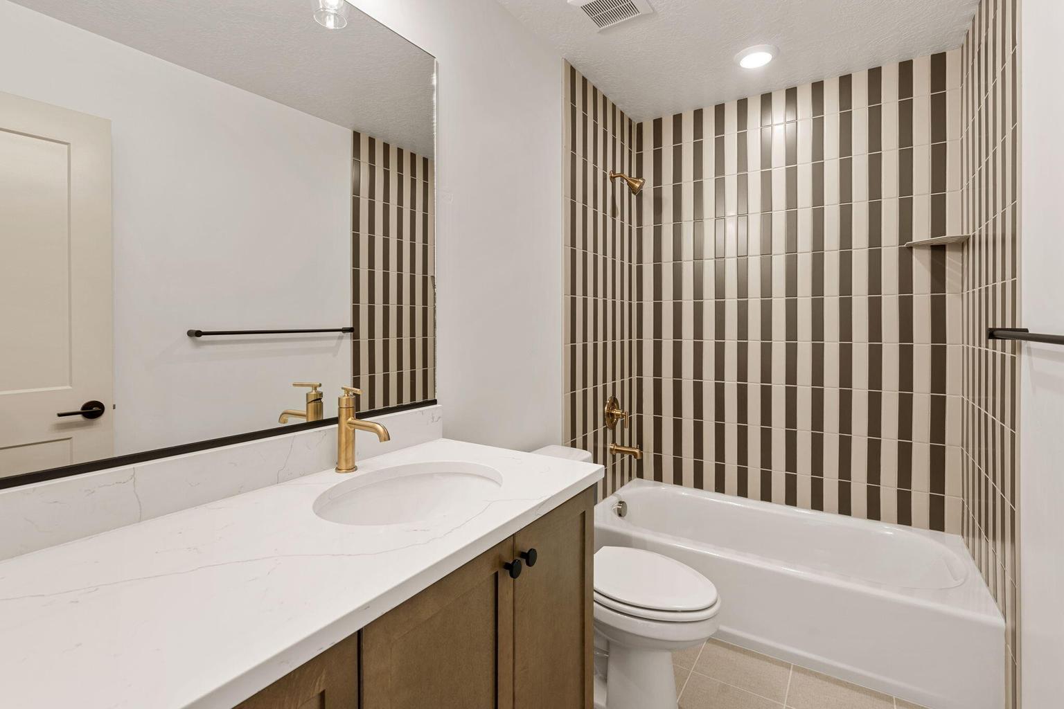 Bathroom featuring a white bathtub with vertically striped wall tile, a single vanity with a white countertop, an undermount sink, and a gold-finish faucet