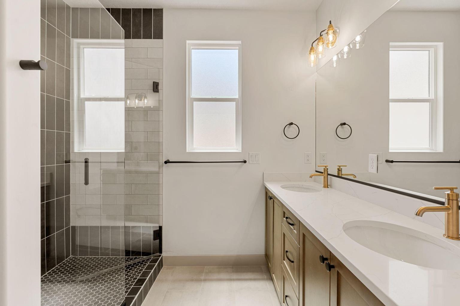 Contemporary bathroom featuring a dual vanity with gold-tone fixtures, a spacious walk-in shower with dark tile surround and mosaic floor, and two frosted windows