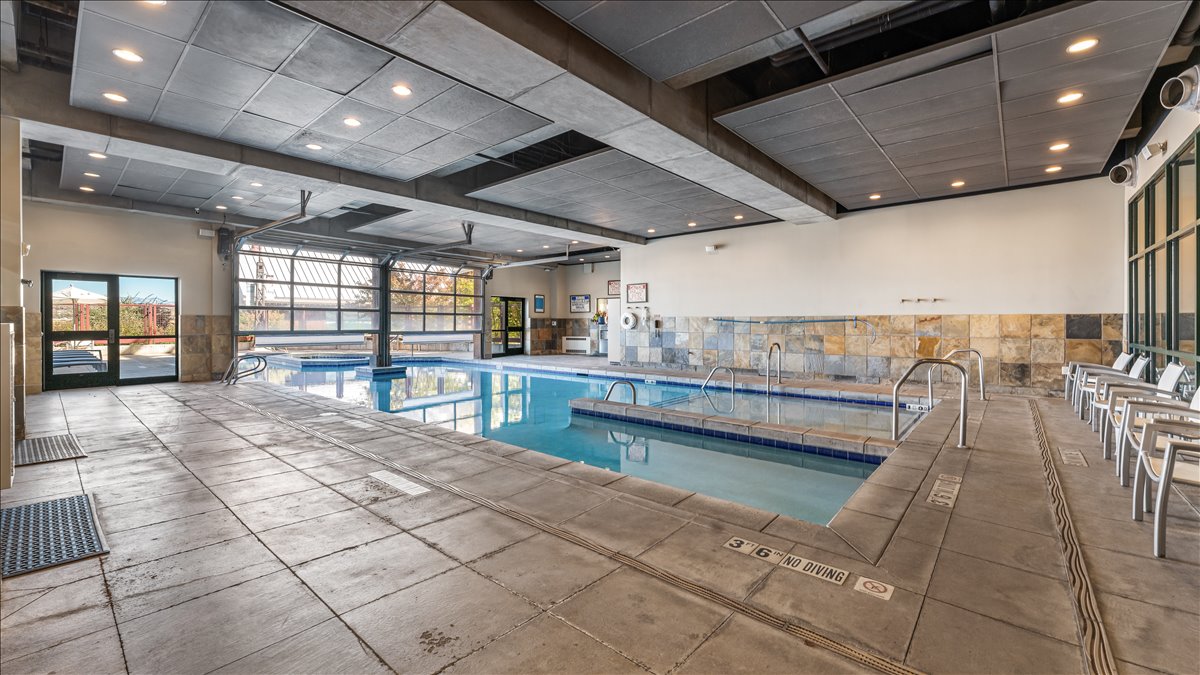 Indoor aquatic facility featuring a lap pool and separate spa, concrete decking, stone wall accents, and an overhead glass garage door