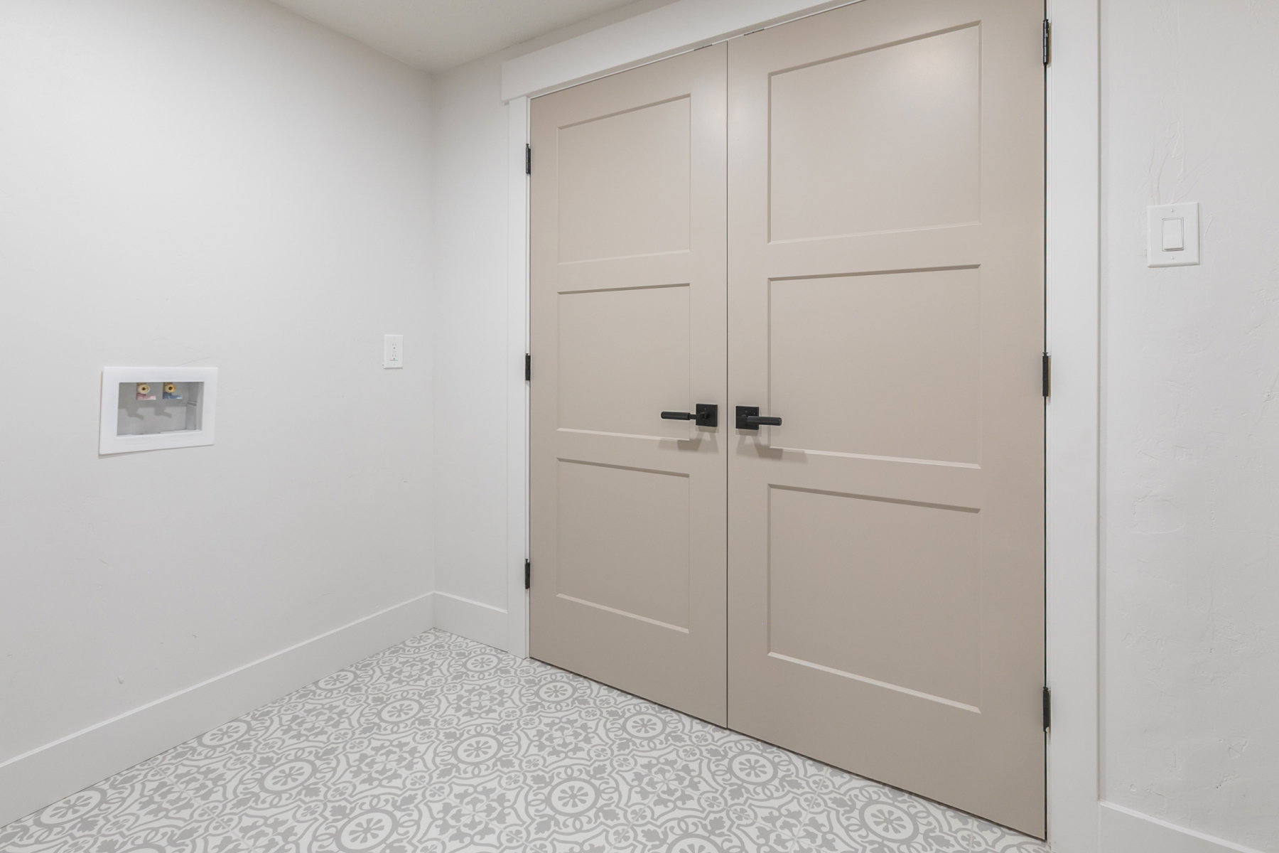 Dual panel doors with matte black hardware, patterned floor tile, white baseboards, and a recessed utility box